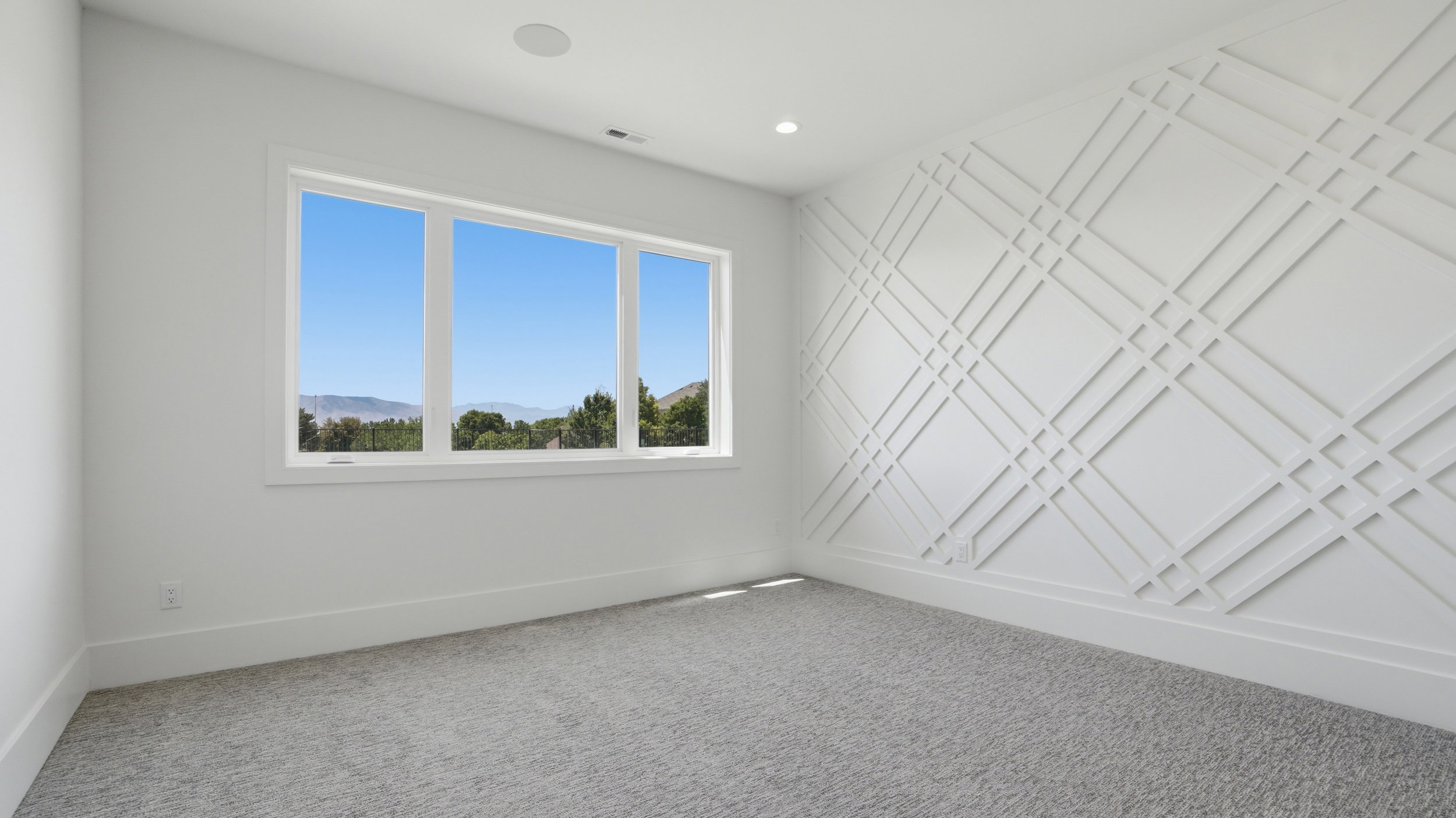 Room featuring a prominent decorative accent wall with geometric paneling, expansive triple-pane window, recessed lighting, ceiling-mounted speaker, and soft grey carpet flooring