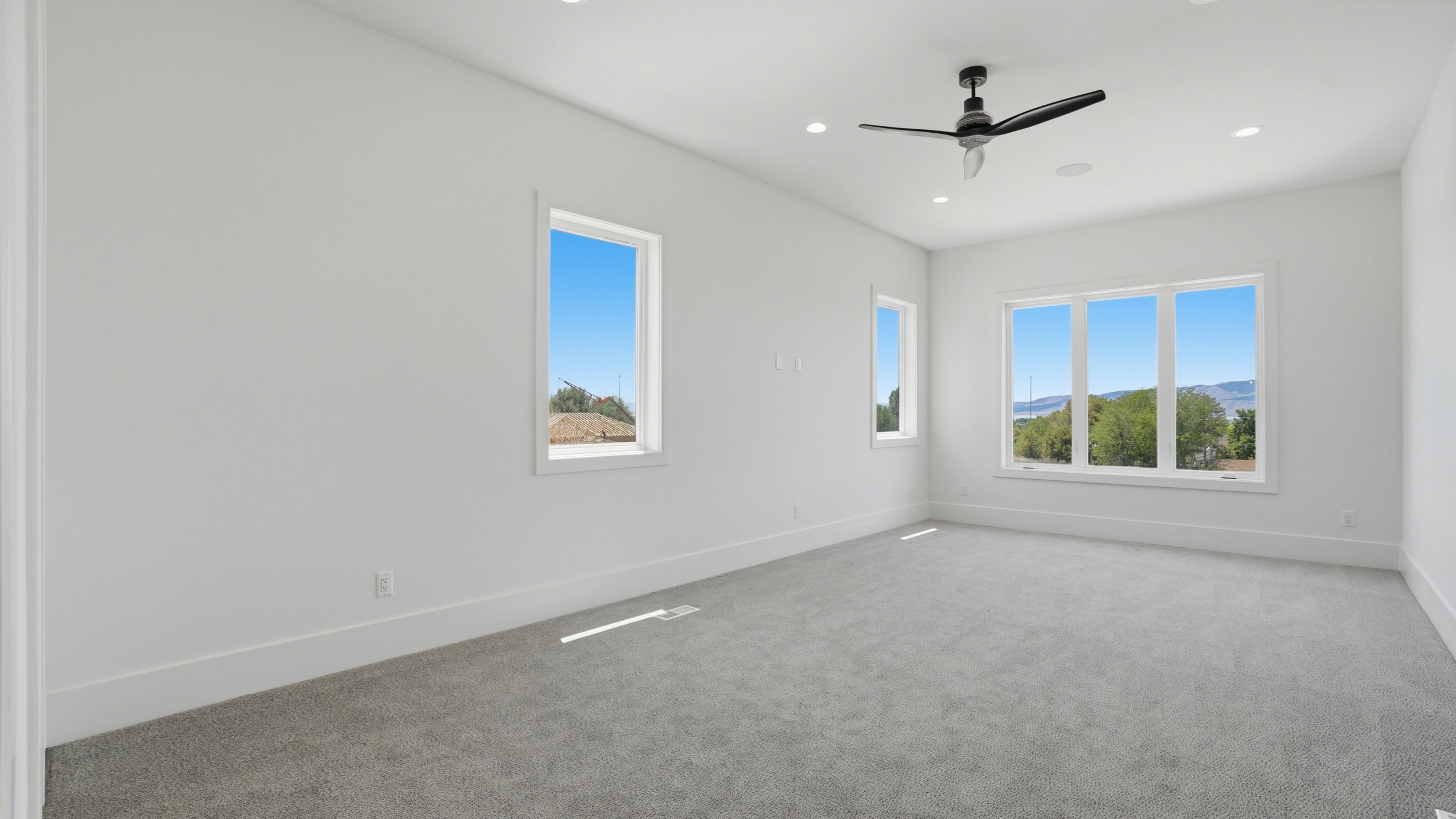 Spacious room featuring light gray carpeting, crisp white walls, recessed ceiling lighting, a modern ceiling fan, and multiple large windows showcasing distant scenery