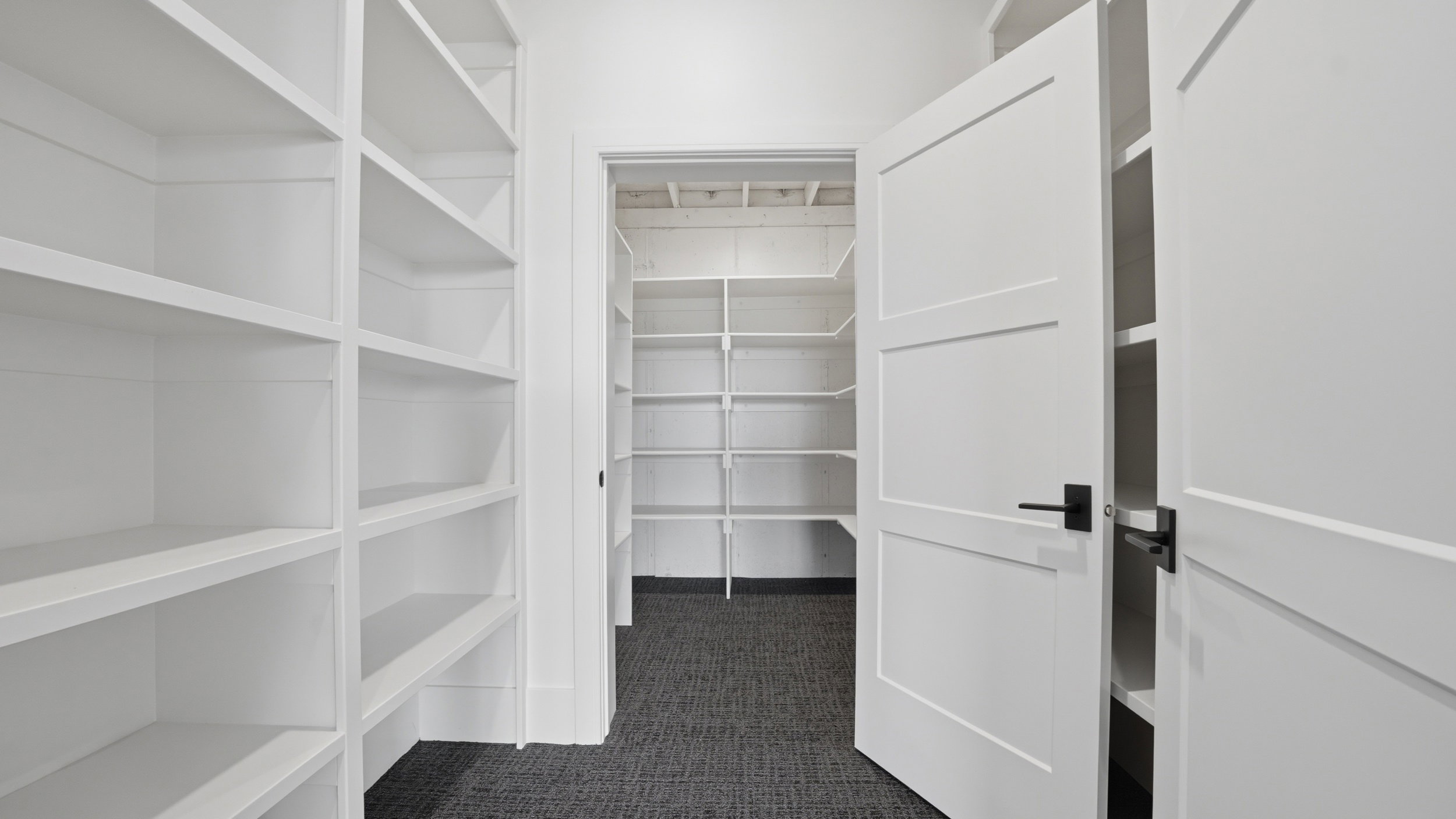 Expansive pantry featuring extensive built-in white shelving, dark gray carpet flooring, and white paneled doors with matte black hardware