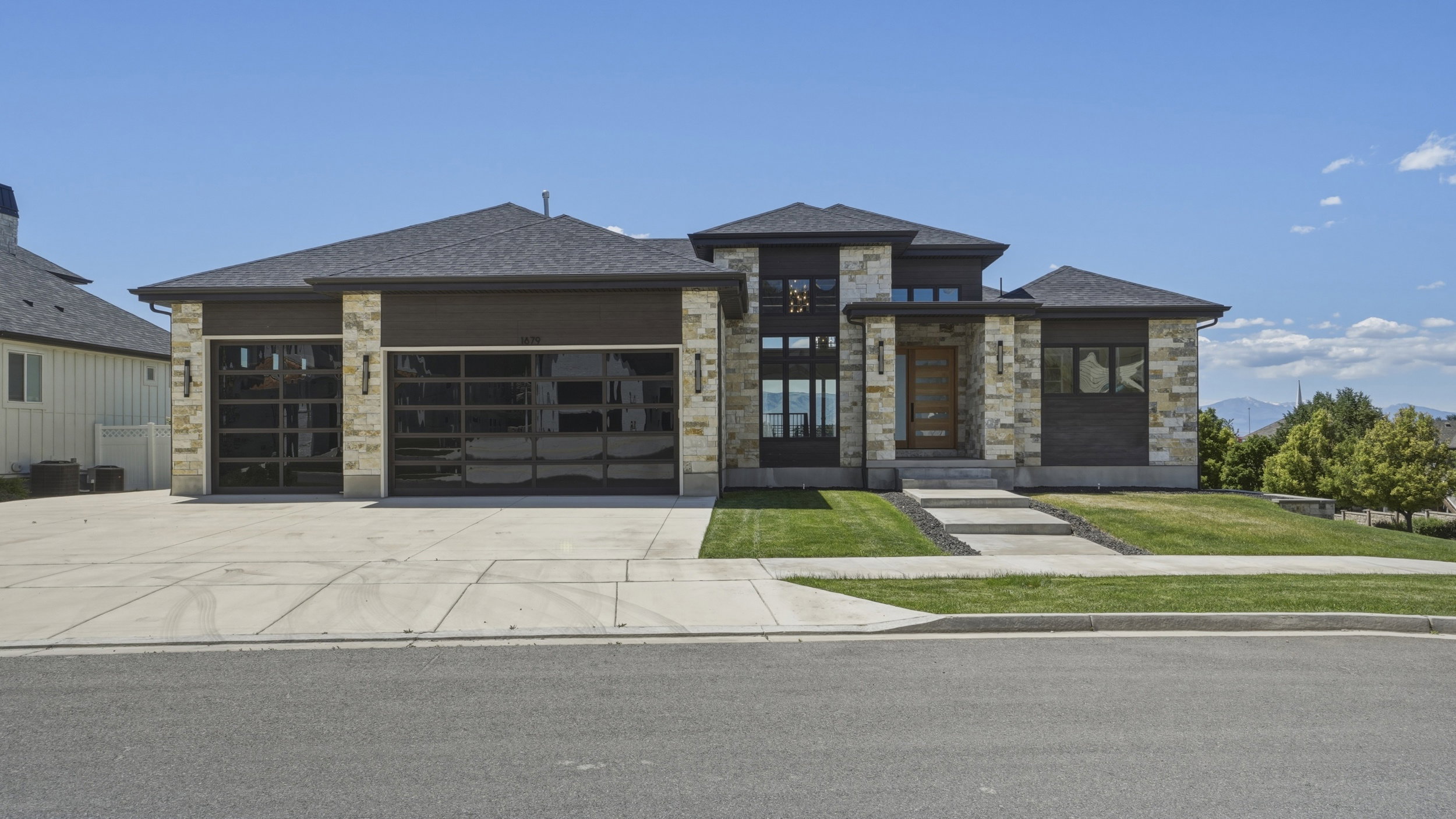 Contemporary residence featuring a stone facade, multi-pane windows, a dark shingle roof, two glass-panel garage doors, and a concrete driveway