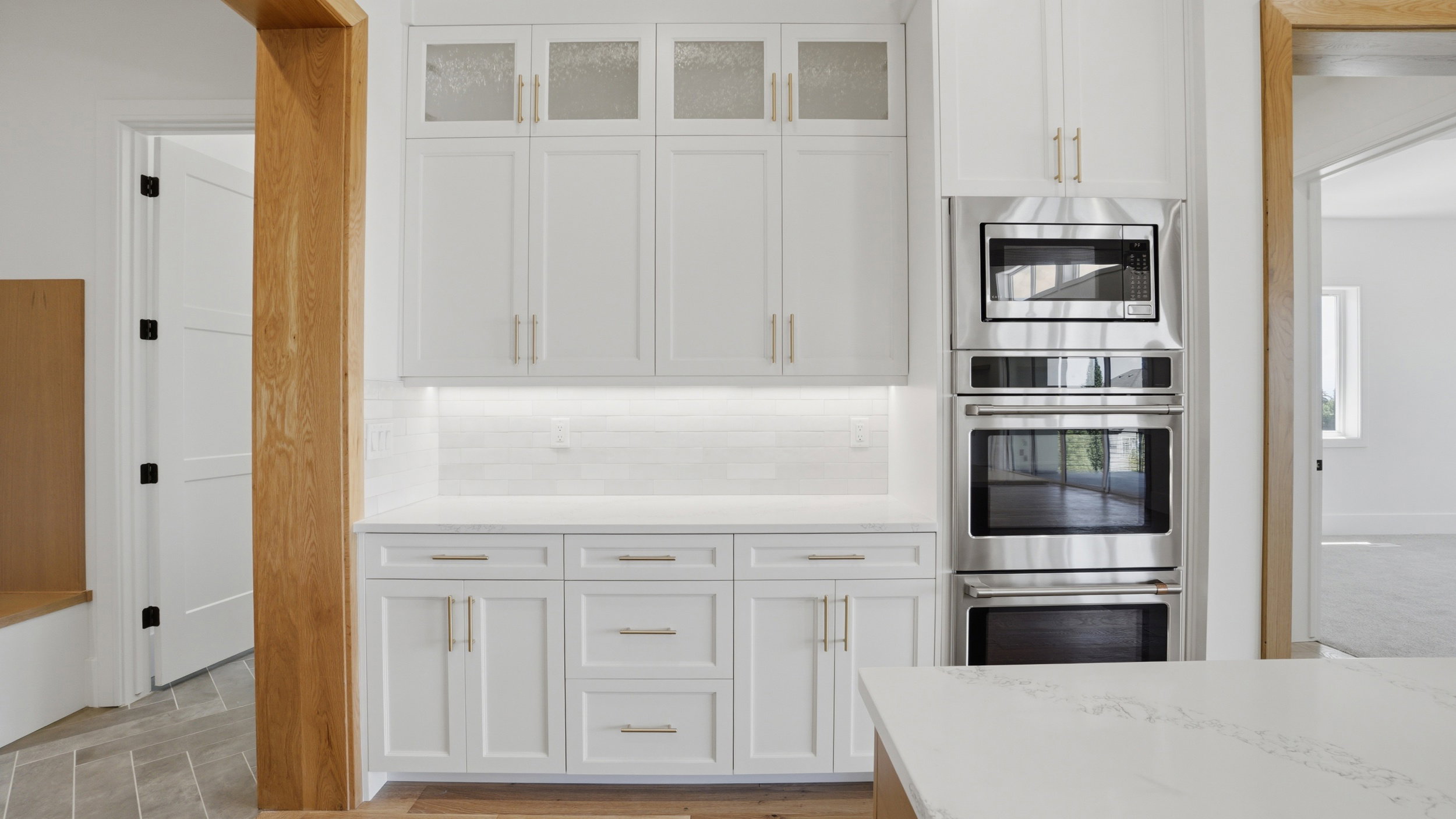 Kitchen featuring white cabinetry with gold-toned hardware, stacked stainless steel appliances, white subway tile backsplash, under-cabinet lighting, and stone countertops