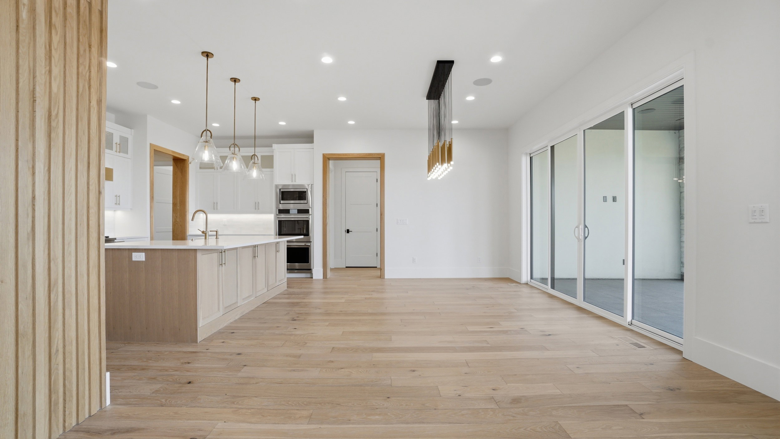 Open-concept interior featuring light wood-finish flooring, a contemporary kitchen with a large island and pendant lighting, white cabinetry, built-in stainless steel appliances, and expansive sliding glass doors