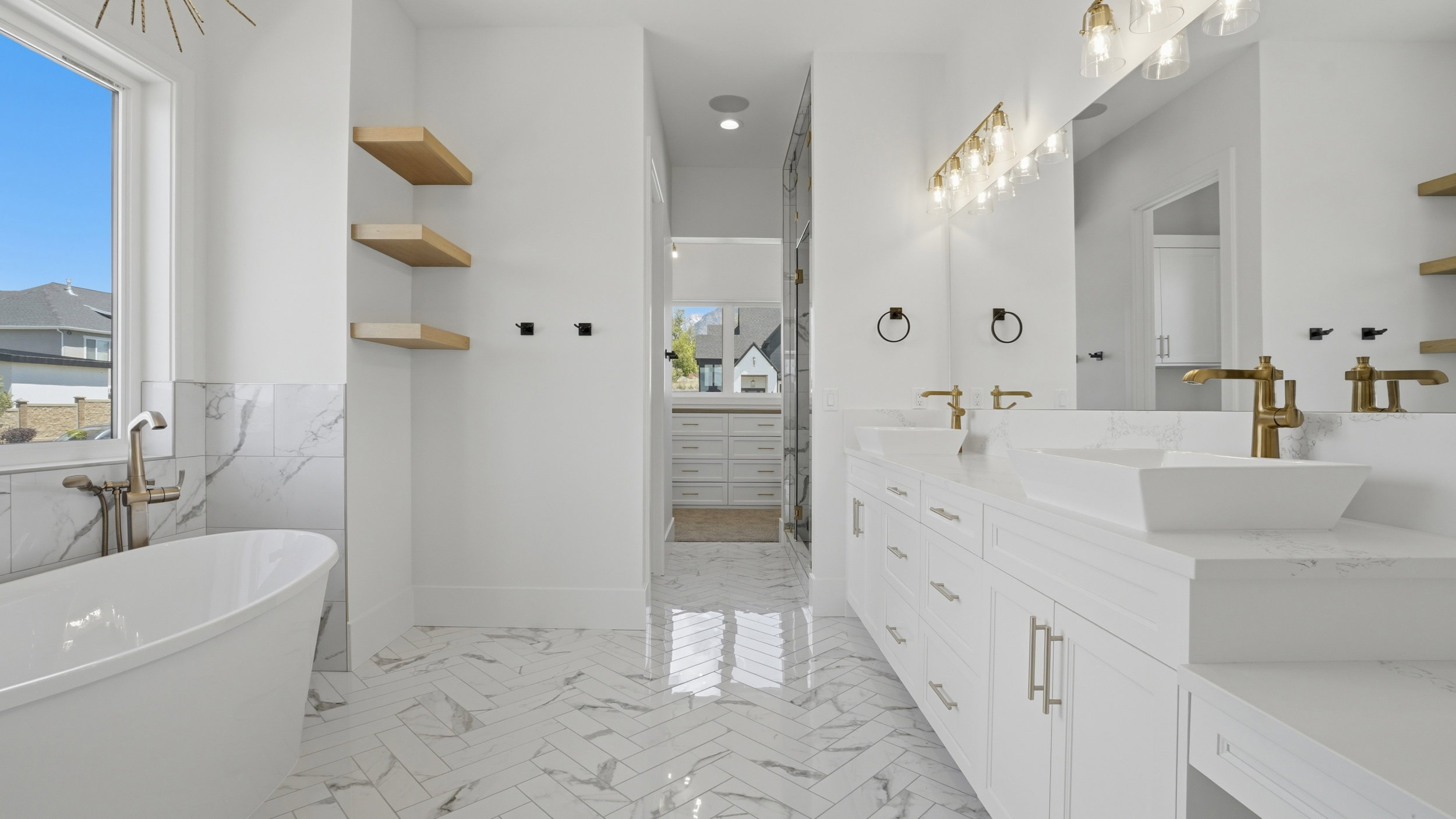 Freestanding soaking tub with brass fixtures, marble-patterned tile, and light wood-finish floating shelves