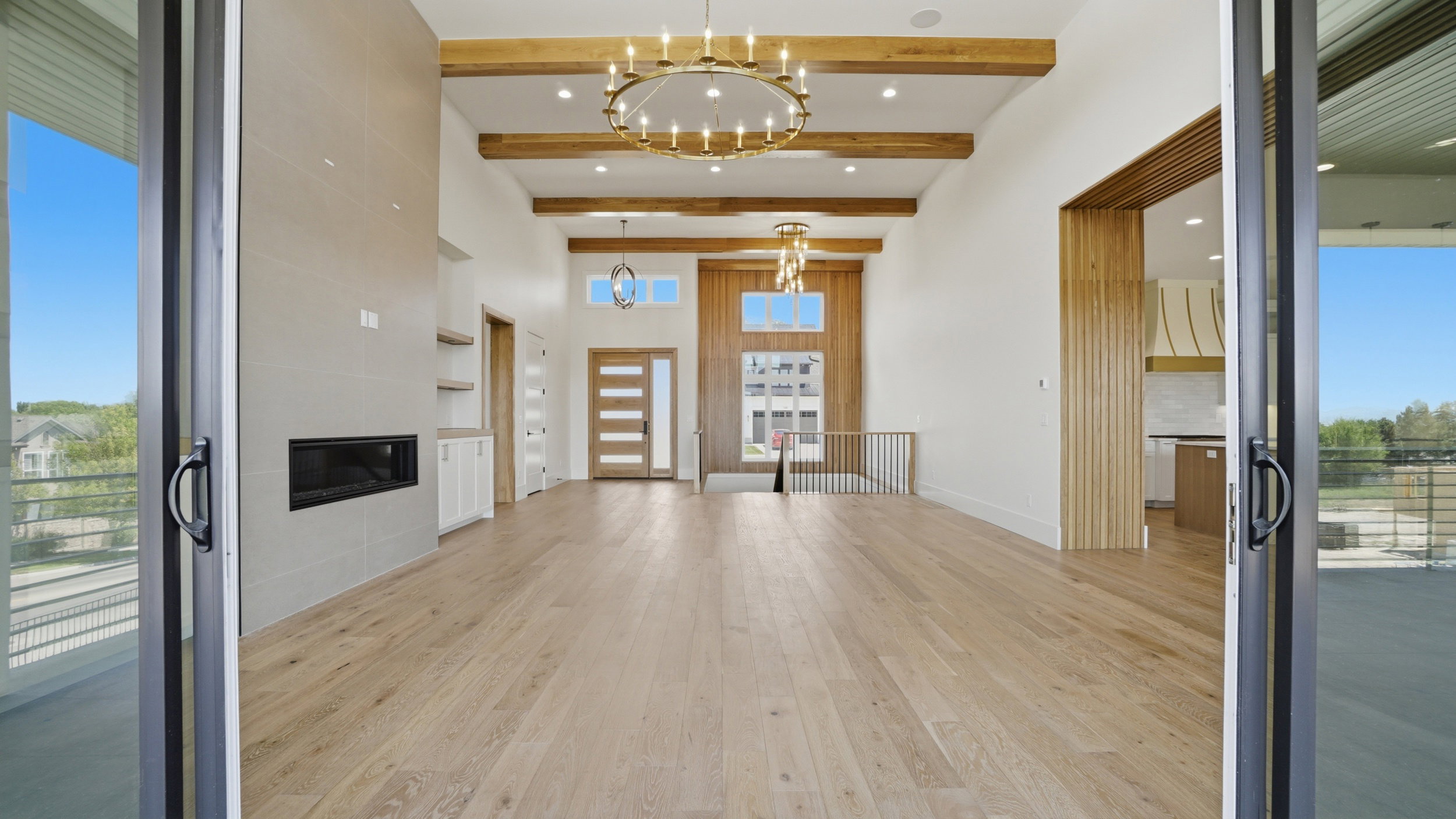 Expansive living space featuring wood-finish flooring, exposed ceiling beams, a linear fireplace set within a tiled accent wall, and contemporary chandeliers