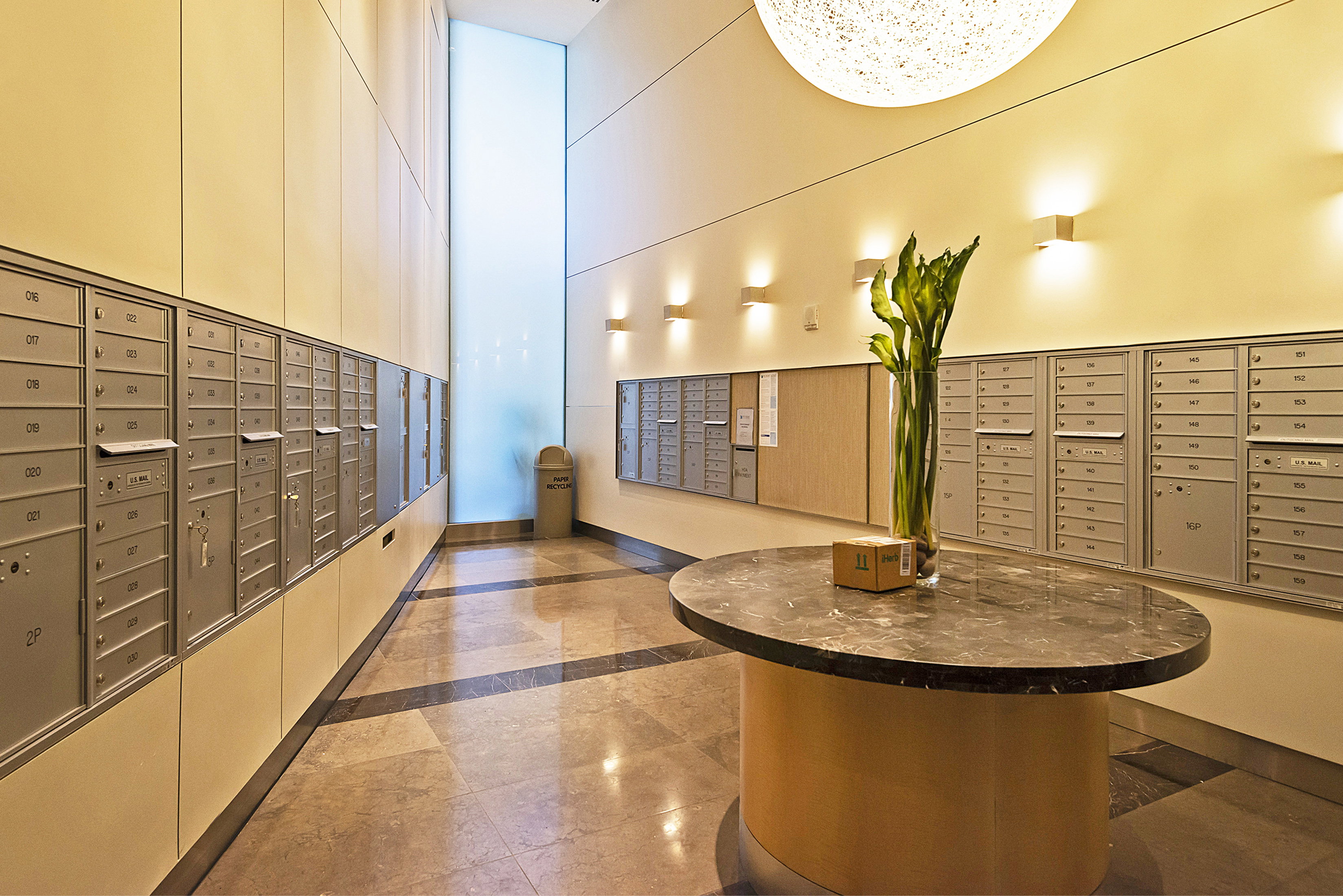 Mailroom featuring extensive wall-mounted mailboxes, a central oval table with a dark stone-finish top, and integrated wall lighting