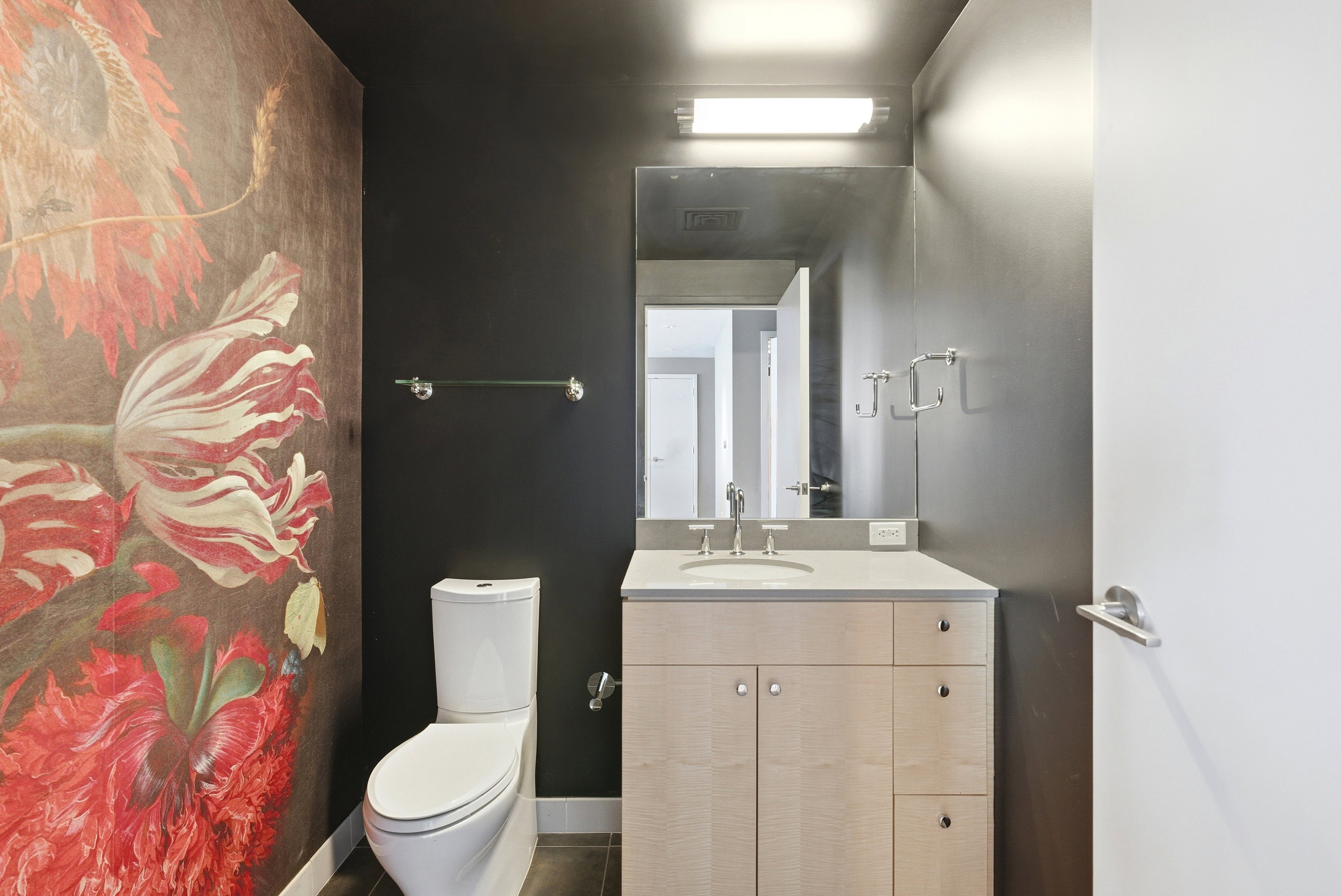 Powder room featuring a large-scale floral accent wall, dark painted walls, a light wood-finish vanity with a light countertop, and a rectangular mirror with integrated lighting
