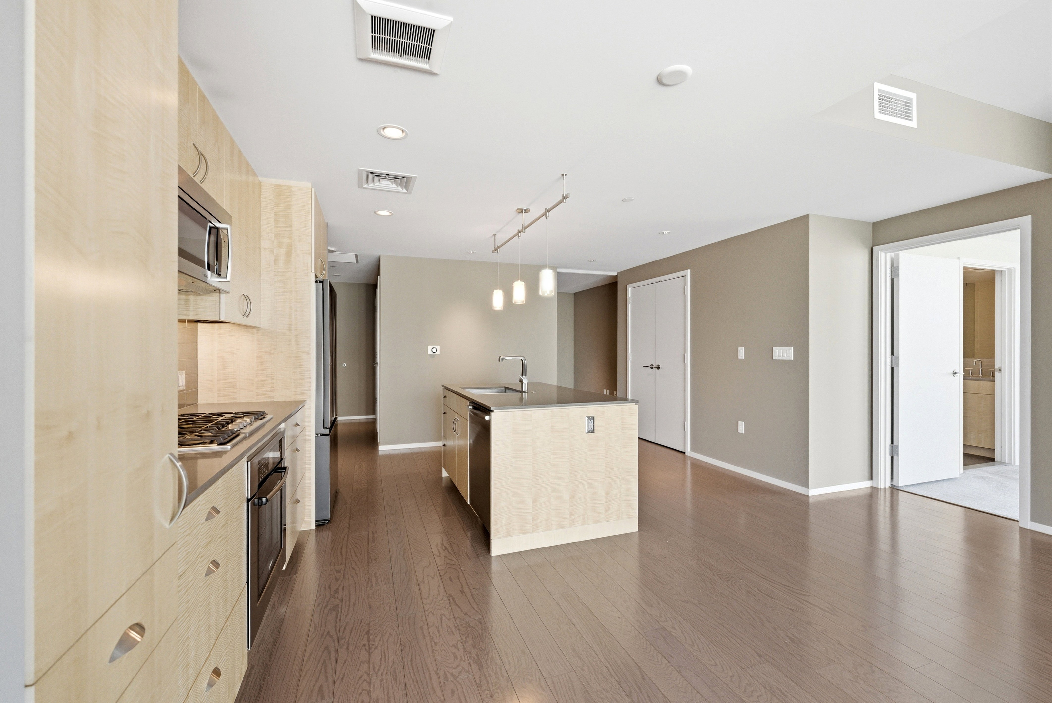 Open-concept kitchen and living area featuring wood-finish flooring, light wood cabinetry, stainless steel appliances, and a central island with a sink and integrated dishwasher