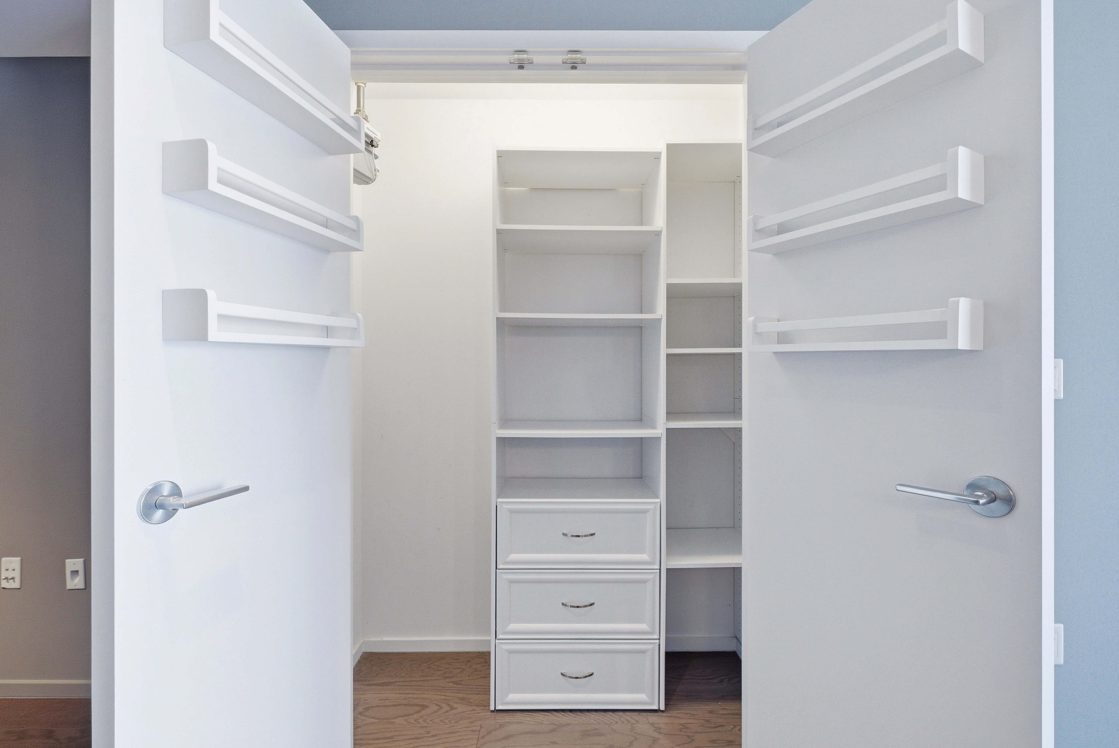 Walk-in closet with custom built-in shelving, multiple storage compartments, three drawers with silver hardware, and wood-finish flooring