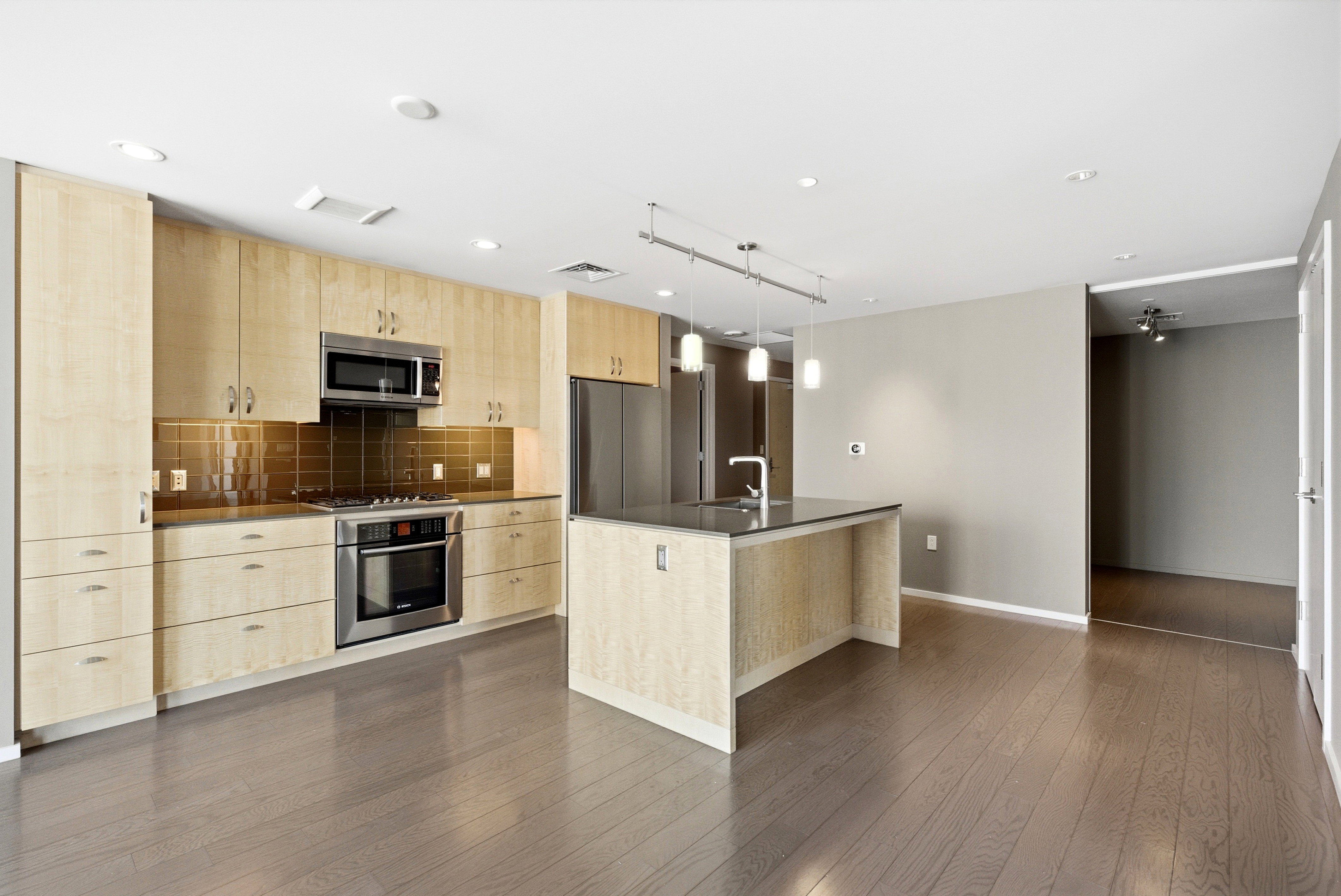 Open-concept kitchen featuring wood-finish flooring, light-toned cabinetry, stainless steel appliances, a center island with a sink, and pendant lighting