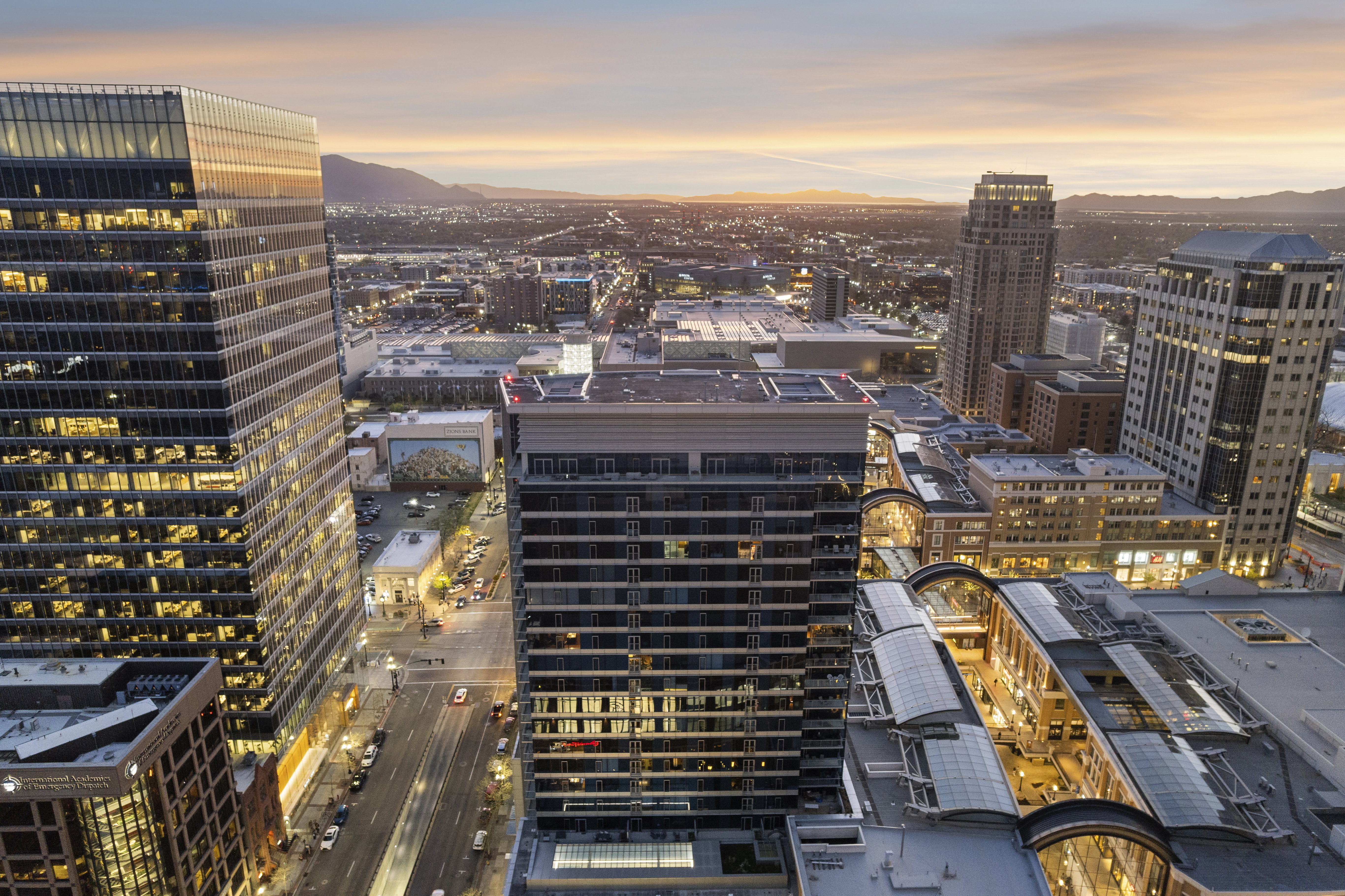 Urban skyline featuring multiple high-rise buildings, a street with streetlights and vehicles, and distant mountains under a sunset sky