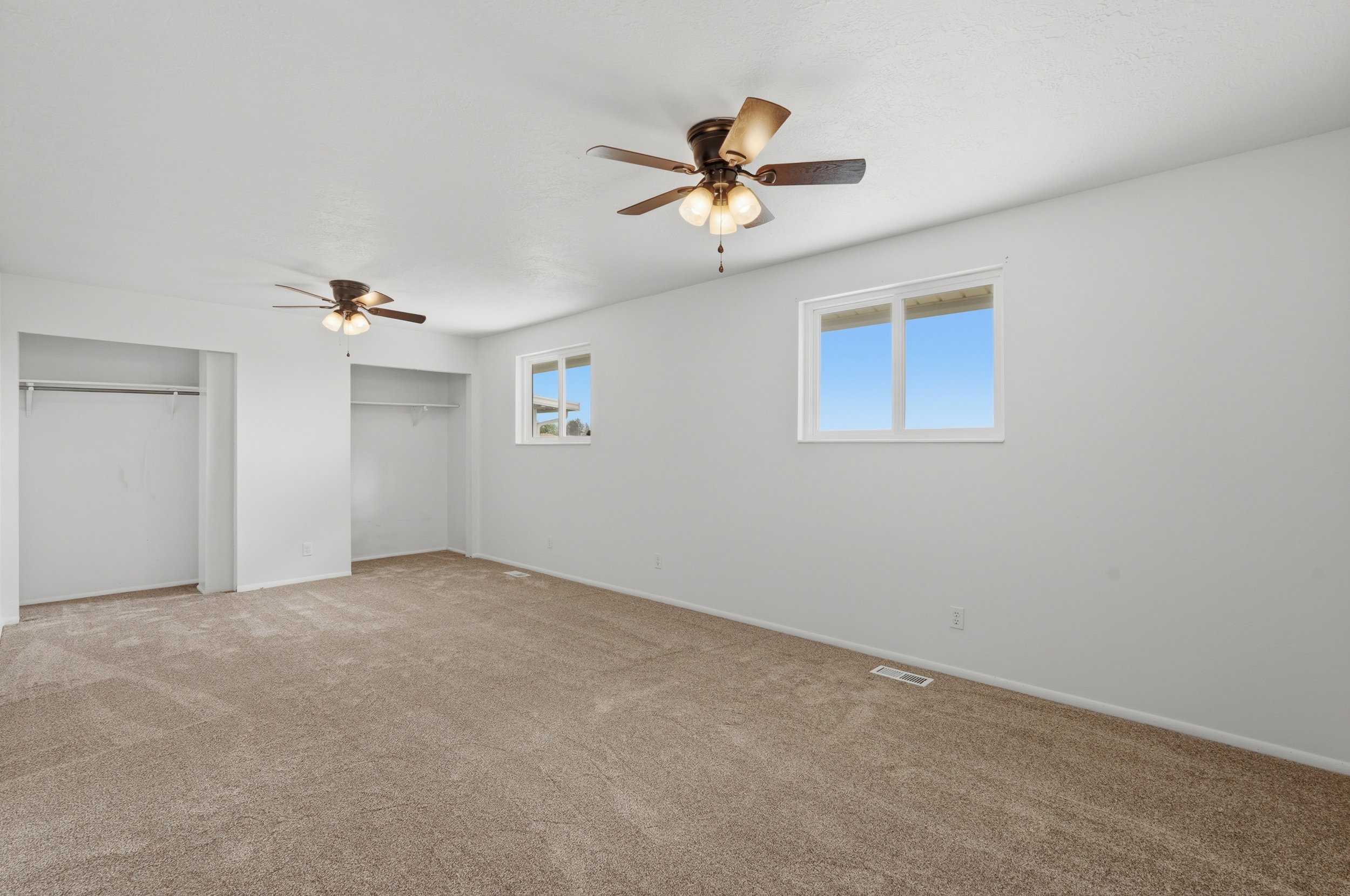 Spacious room featuring neutral carpeting, two ceiling fans with integrated lighting, two windows, and two open closet areas with hanging rods