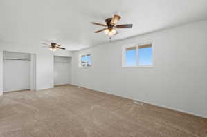 Spacious room featuring neutral carpeting, two ceiling fans with integrated lighting, two windows, and two open closet areas with hanging rods