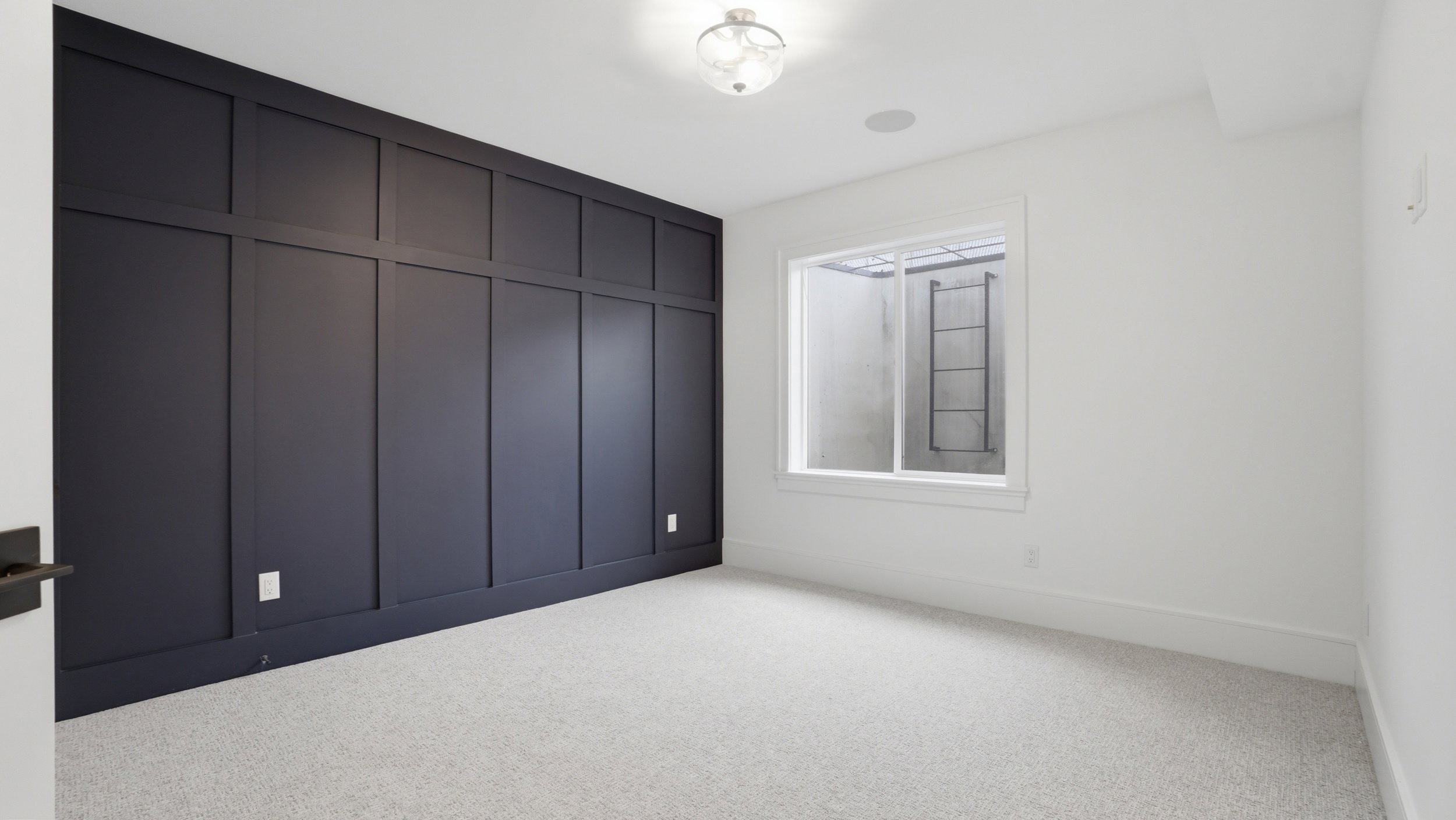 Accent wall with dark wainscoting and trim, light gray textured carpet, recessed ceiling speaker, ceiling-mounted light fixture, and white window trim