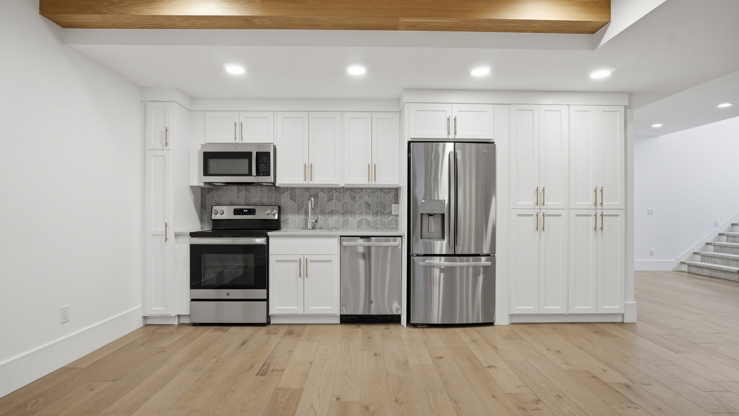 Kitchenette featuring stainless steel appliances, white cabinetry with gold-tone hardware, a gray herringbone tile backsplash, and light wood-finish flooring