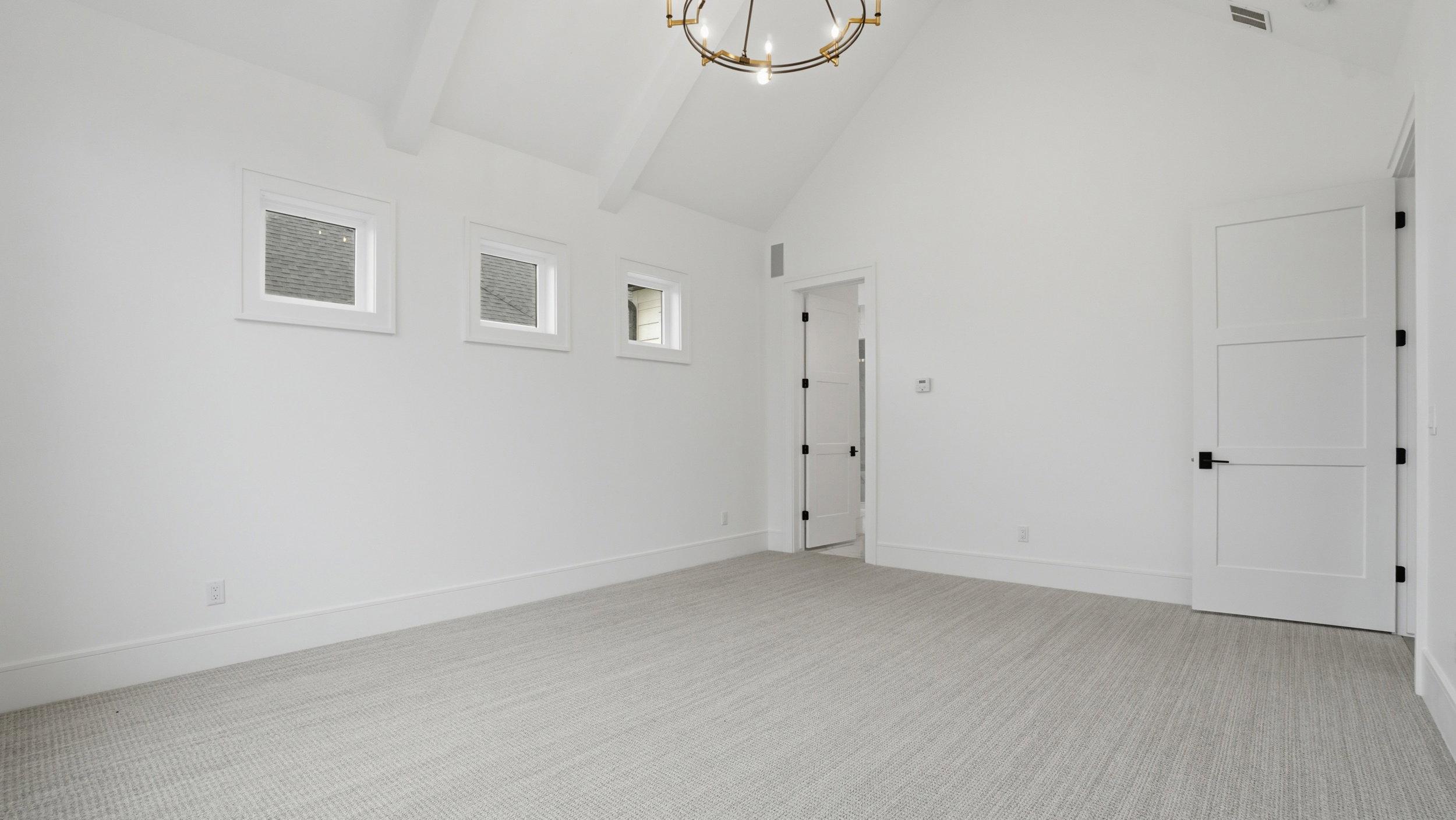 Spacious room featuring vaulted ceilings with exposed beams, light-colored carpeting, multiple windows, and a modern chandelier