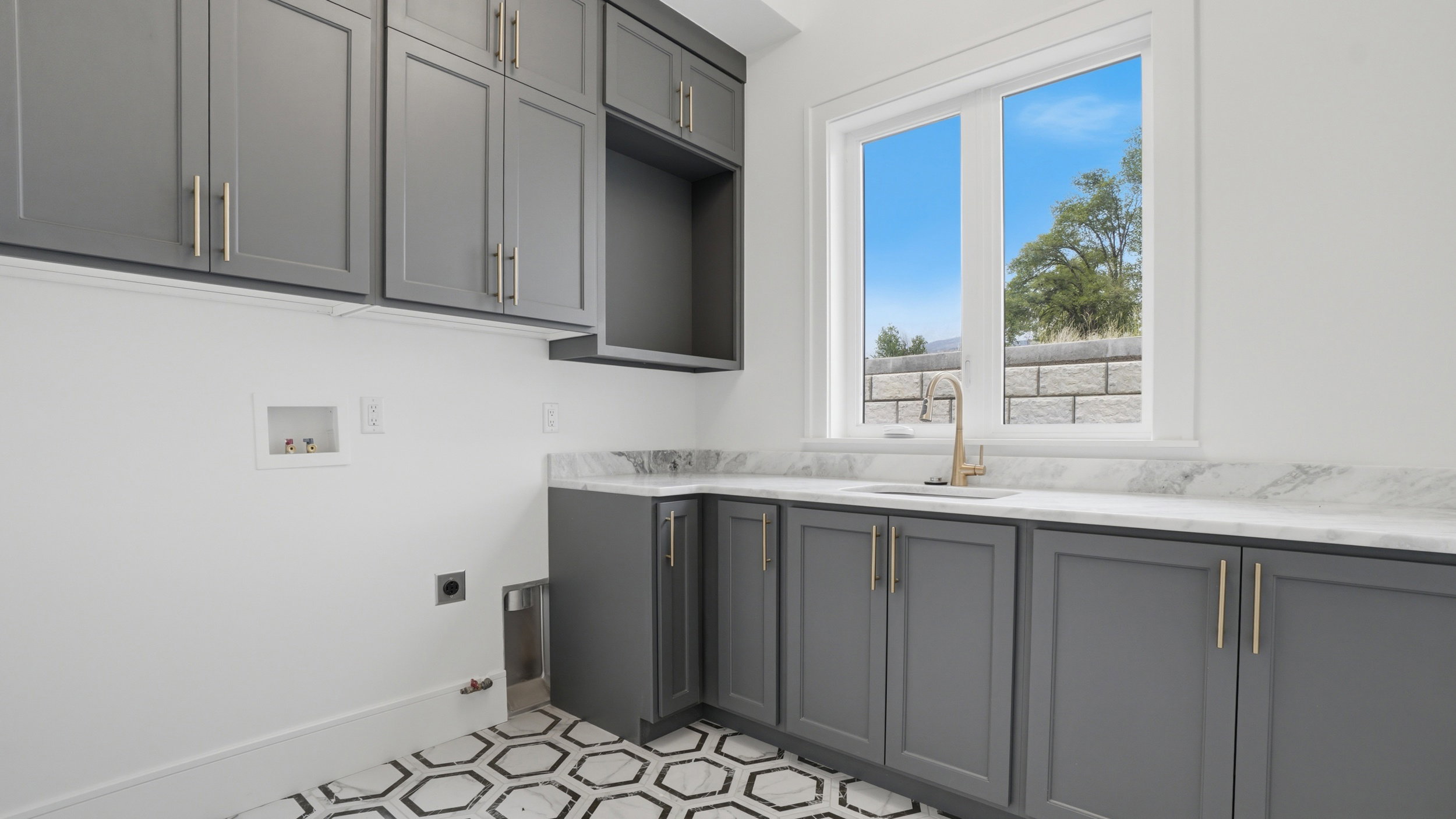 Laundry room featuring gray cabinetry, a white countertop, an undermount sink with a gold-tone faucet, and hexagonal floor tiling