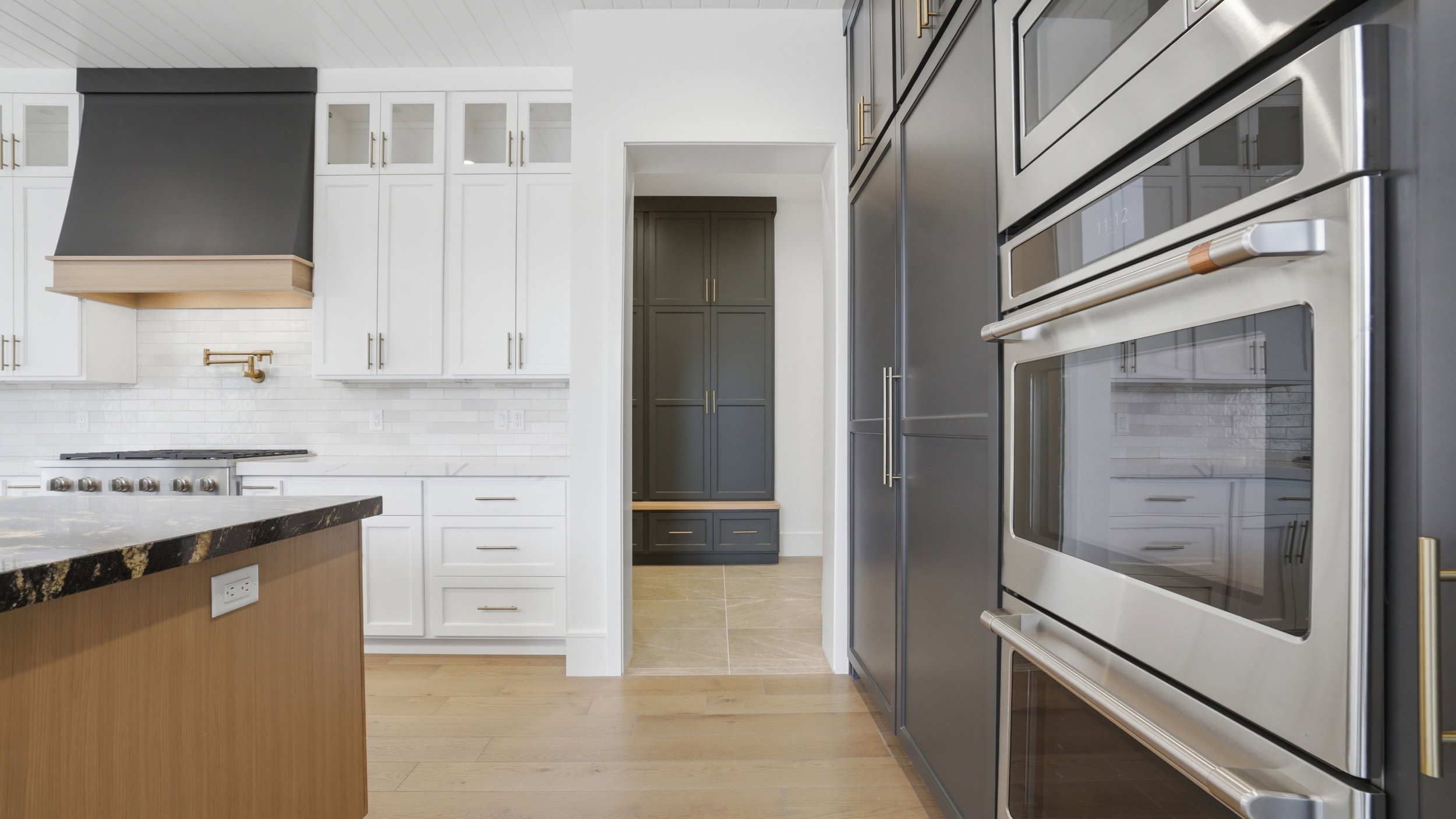 Gourmet kitchen featuring a built-in double oven, full-height dark cabinetry, a light wood-finish island with a dark countertop, white upper and lower cabinetry, and a black range hood with a wood-finish accent