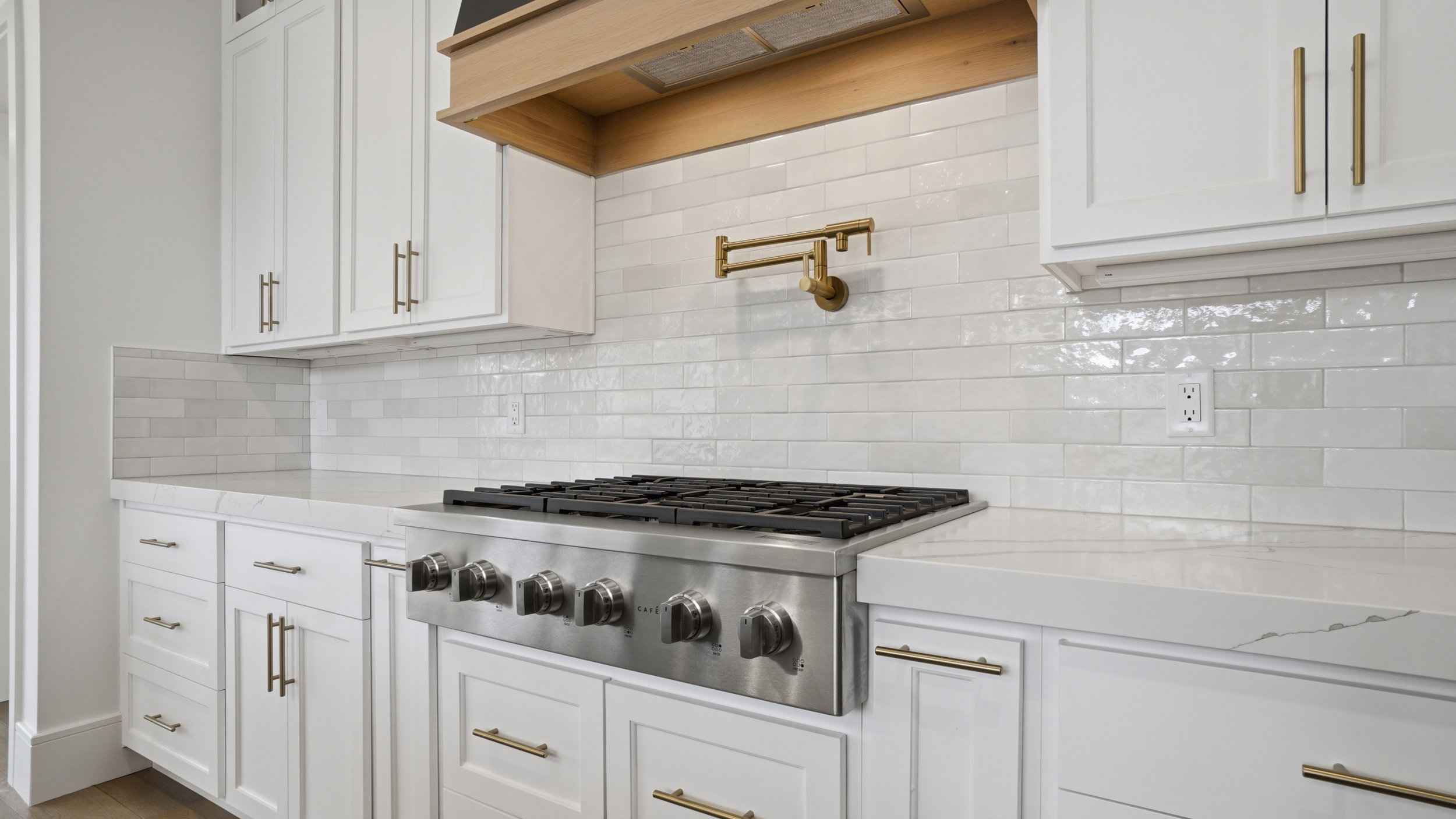 Gourmet kitchen featuring a stainless steel range, a brass pot filler, white shaker-style cabinetry with brass hardware, a light wood-finish range hood, and a white subway tile backsplash