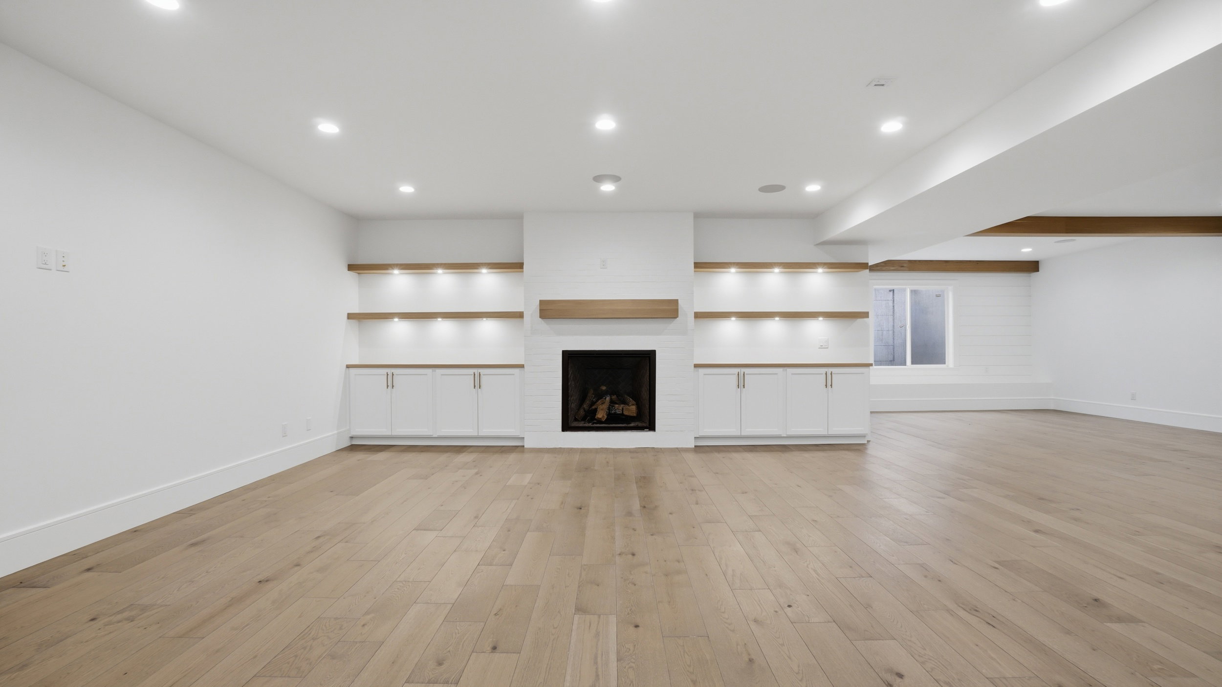 Spacious room featuring light wood-finish flooring, a contemporary fireplace with a wood mantle, and built-in cabinetry with integrated shelving and lighting