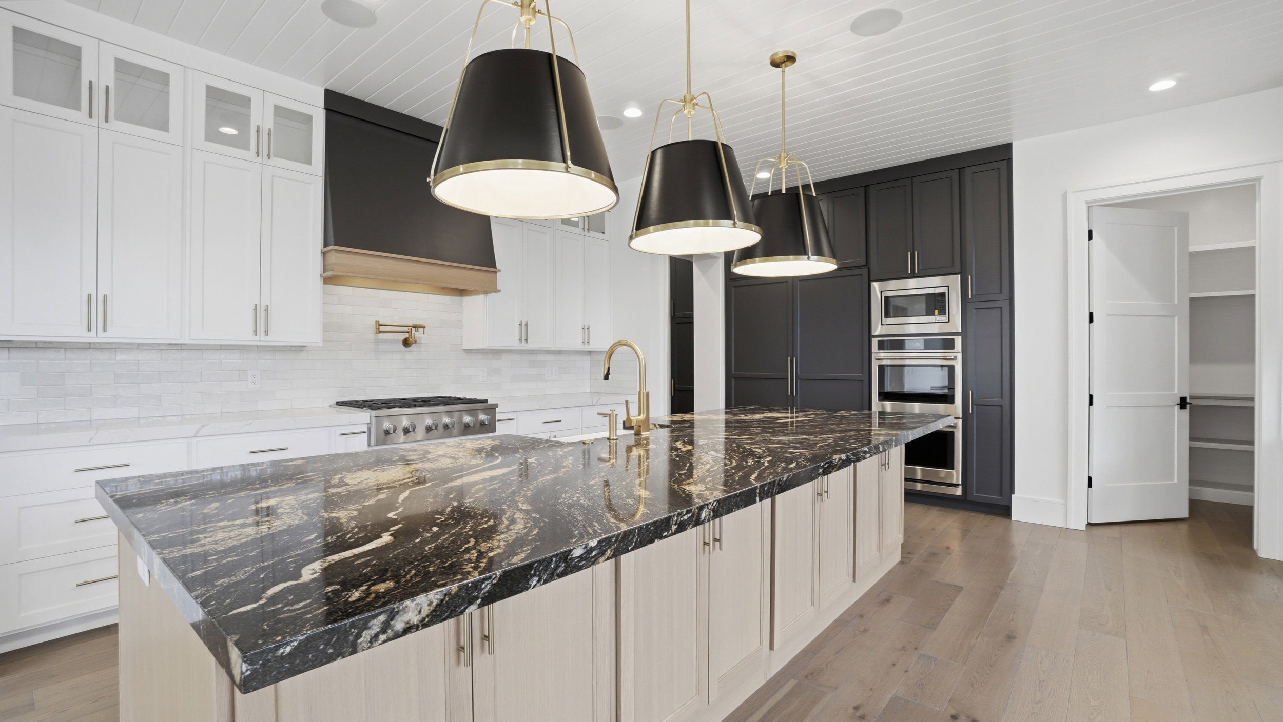 Kitchen featuring a large island with a patterned countertop, brass-toned fixtures, white shaker-style cabinetry, dark cabinetry surrounding built-in appliances, and light wood-finish flooring