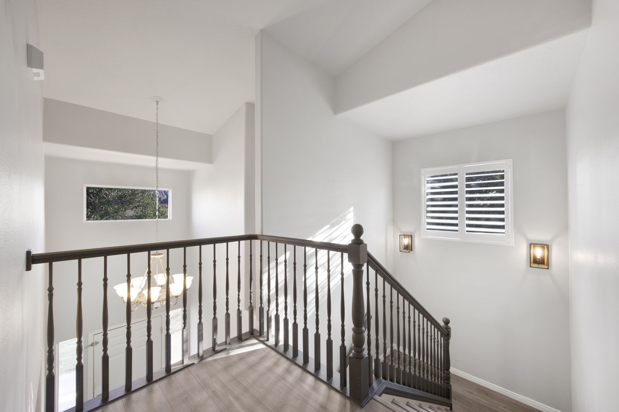 Stairwell featuring a dark wood newel post and balusters, wood-finish flooring, two wall-mounted sconces, and a window with plantation shutters