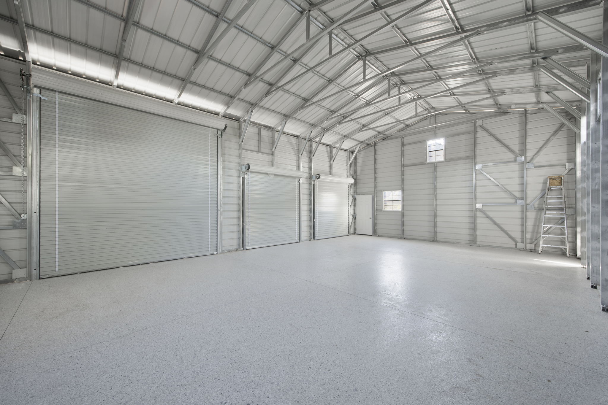 Spacious interior featuring a high arched ceiling with exposed metal truss work, polished concrete flooring, three large roll-up doors, and several windows providing natural light