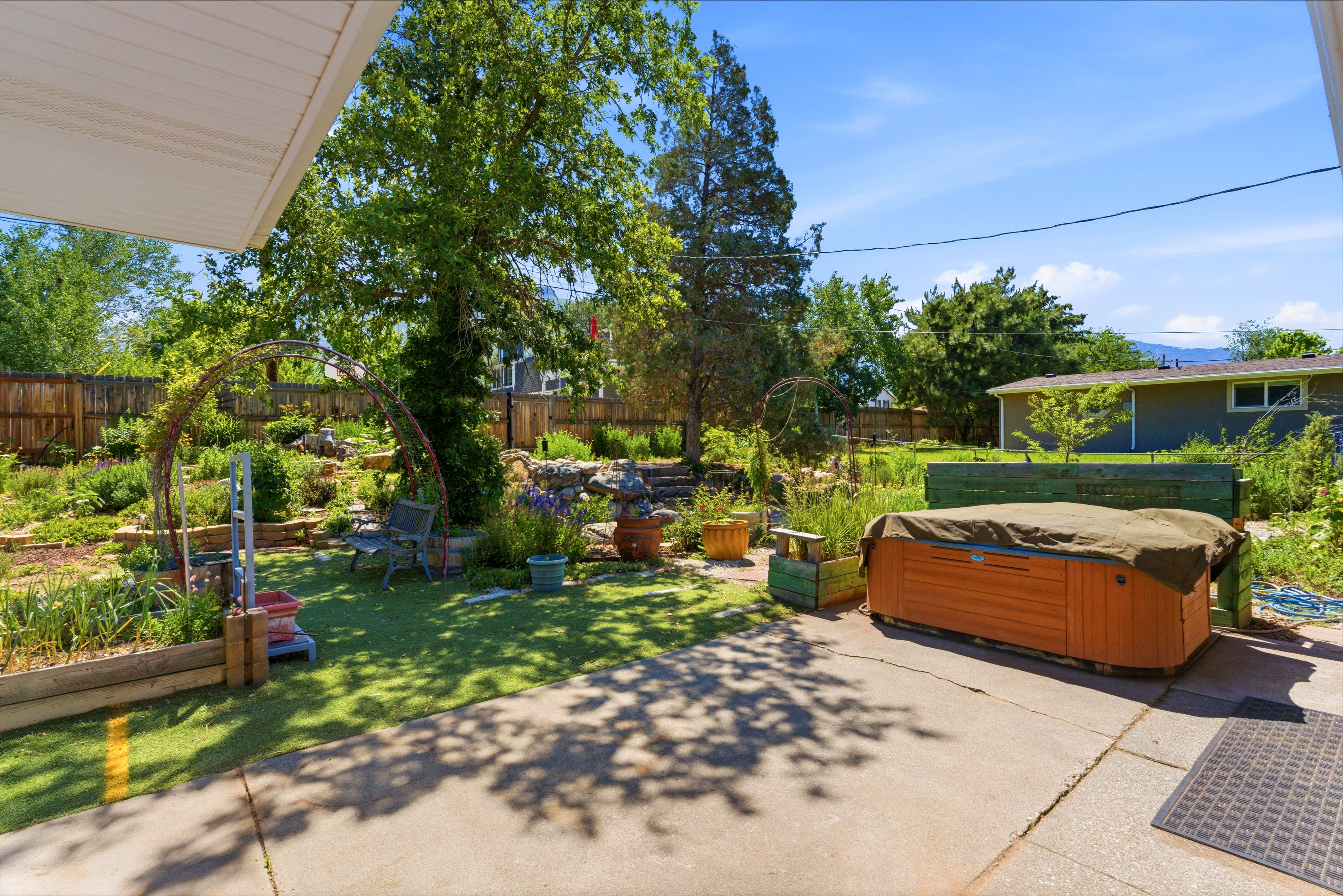 Expansive backyard featuring a concrete patio, a covered spa, a tiered garden with natural rock accents, mature trees, and a full privacy fence
