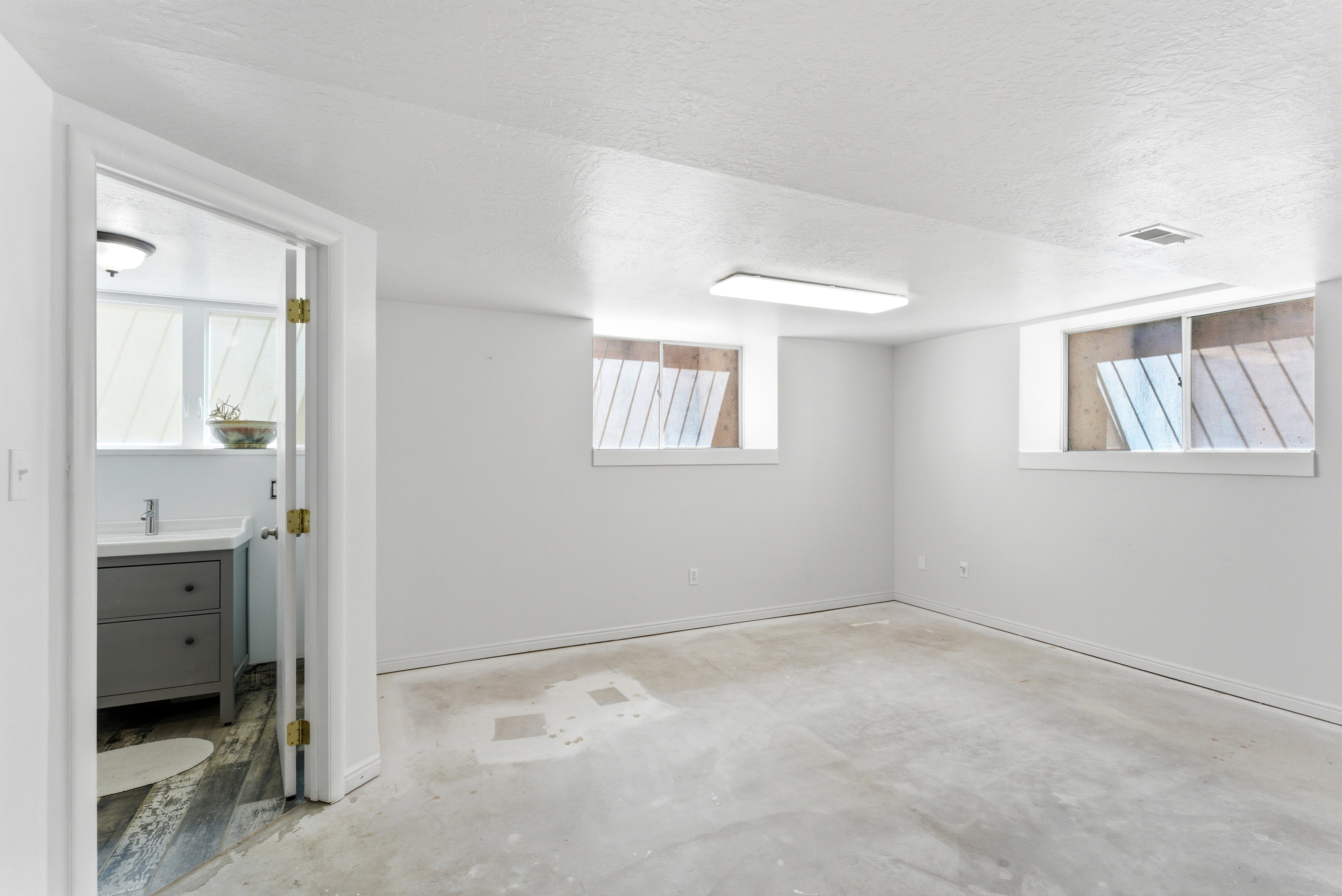 Spacious room featuring two recessed windows, a rectangular ceiling light fixture, and a concrete floor, with an adjacent room showcasing a single vanity with a white countertop
