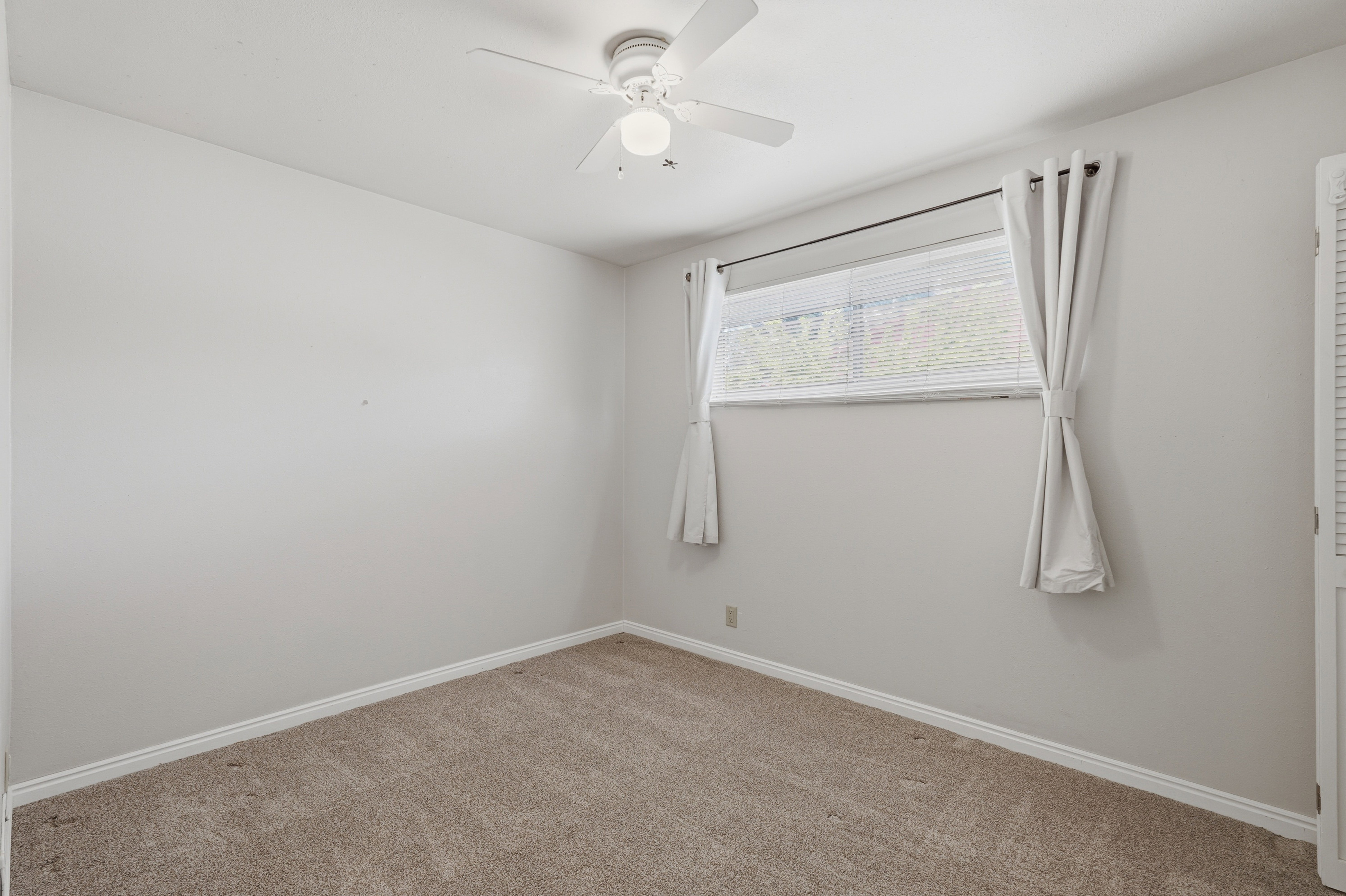 Neutral-toned room featuring light beige carpeting, white baseboards, and a window with white blinds and drapes
