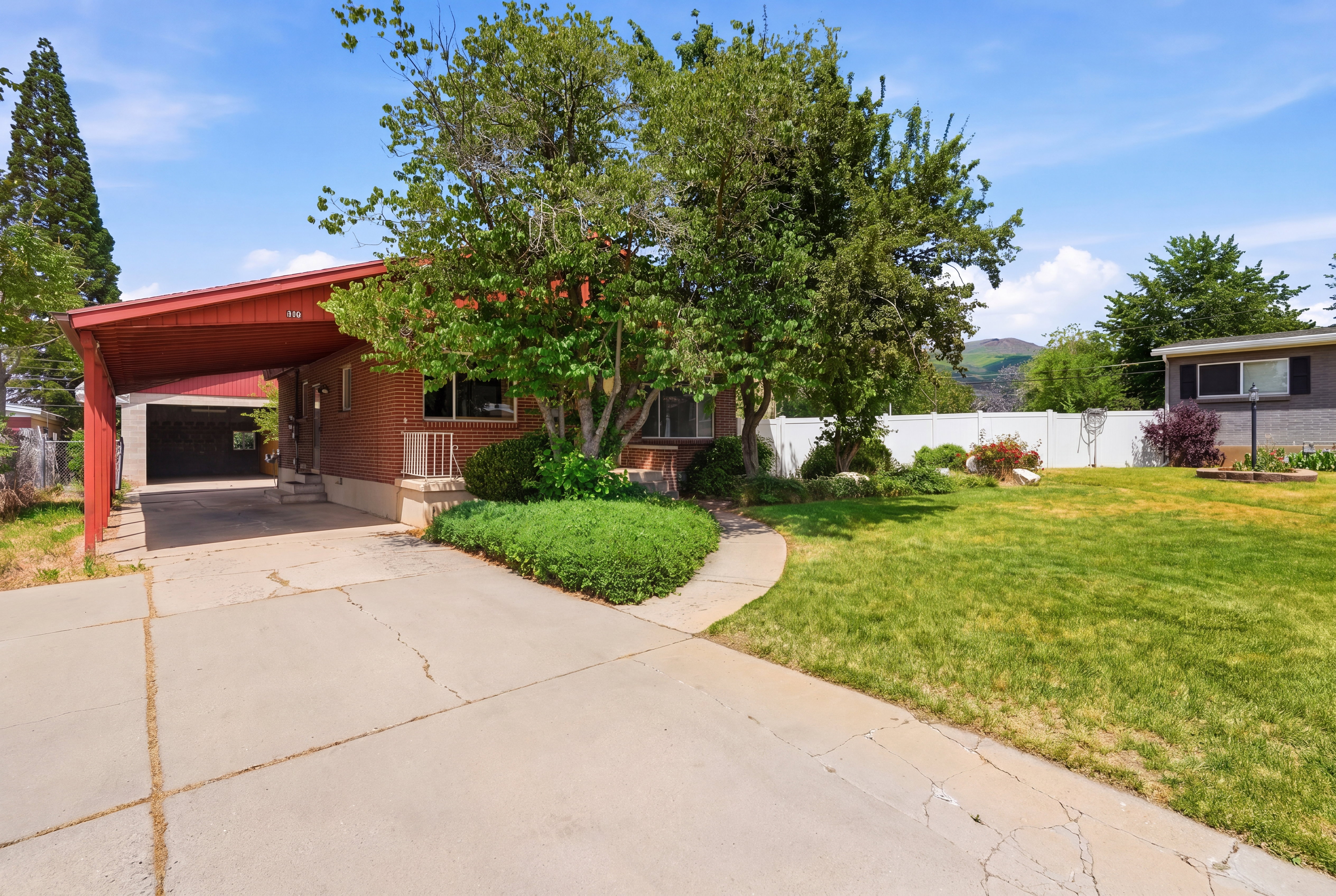 Brick exterior with a red-toned carport, established front yard landscaping, and a concrete driveway