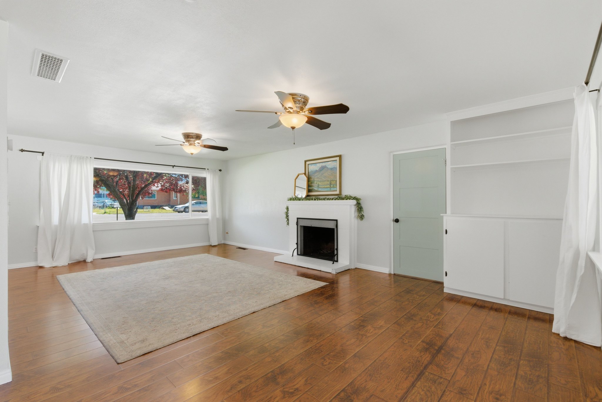 Spacious room featuring wood-finish flooring, a prominent fireplace with a white mantel, and a large picture window