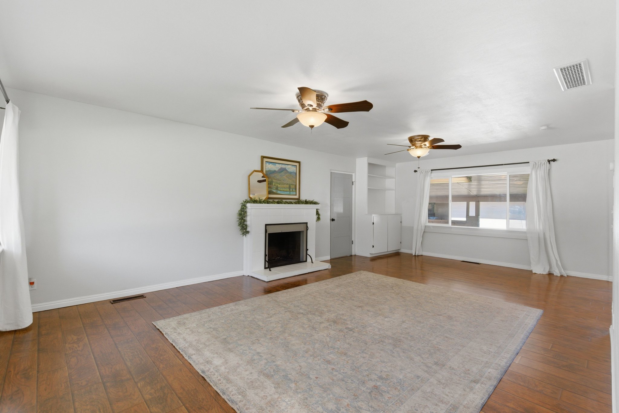 Hardwood-style flooring throughout a spacious room featuring a white brick fireplace, built-in shelving, and two ceiling fans