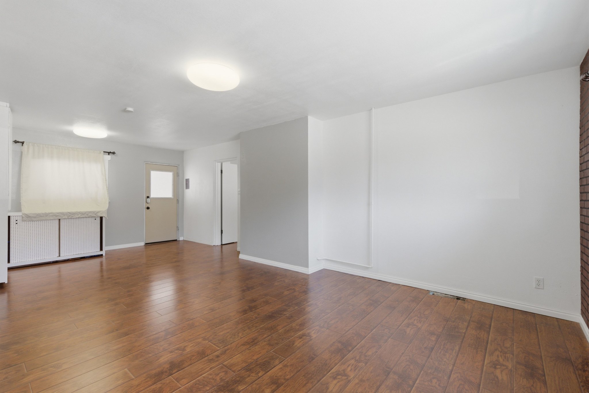 Spacious room with wood-finish flooring, light grey walls, white baseboards, and a partial exposed brick wall accent