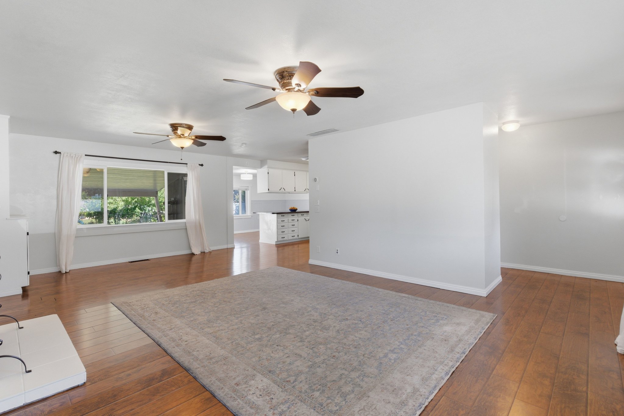Spacious interior with wood-finish flooring, two ceiling fans with integrated lighting, a large window, and a partial wall separating spaces