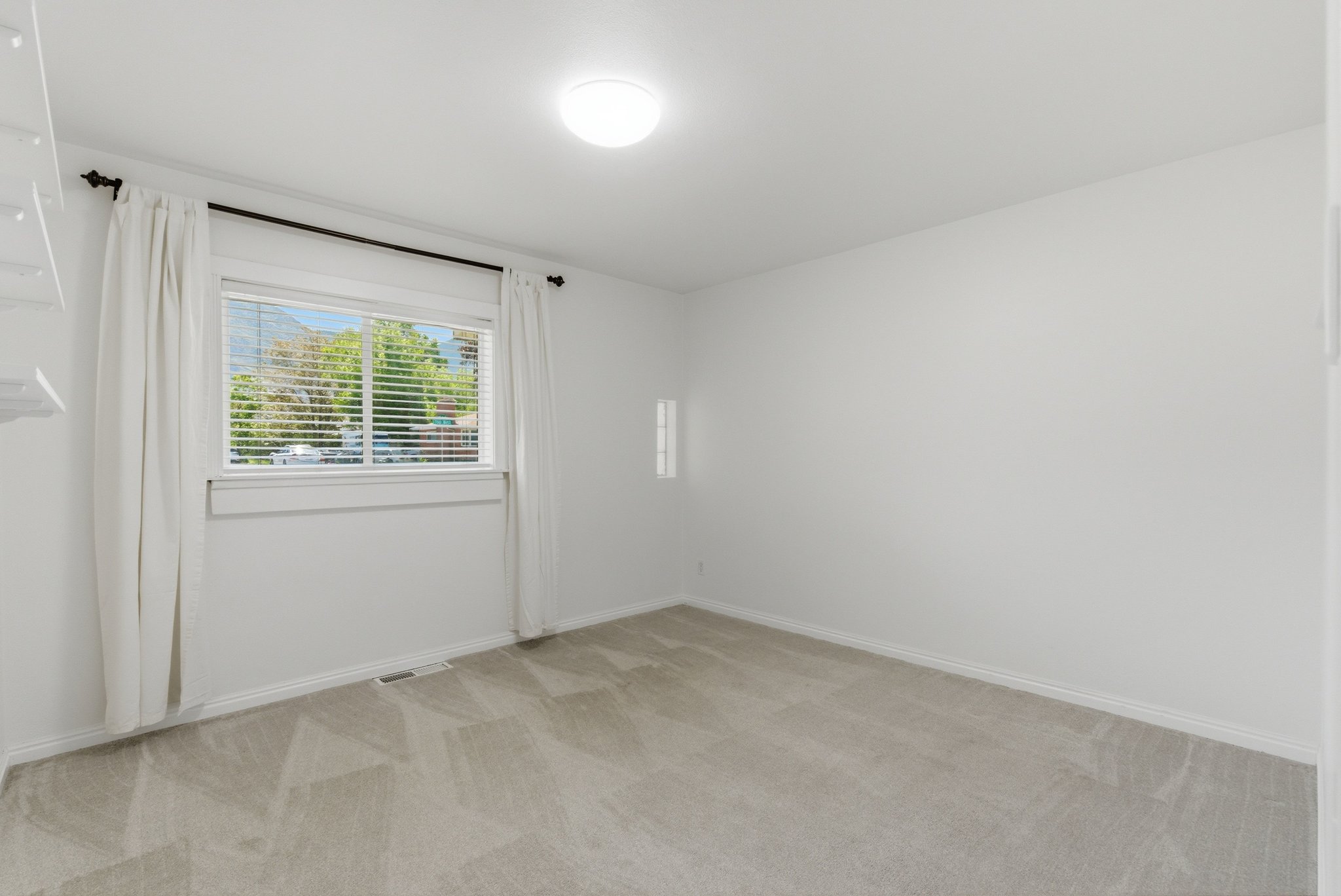 Carpeted room with a ceiling-mounted light fixture and a window featuring horizontal blinds and white drapes