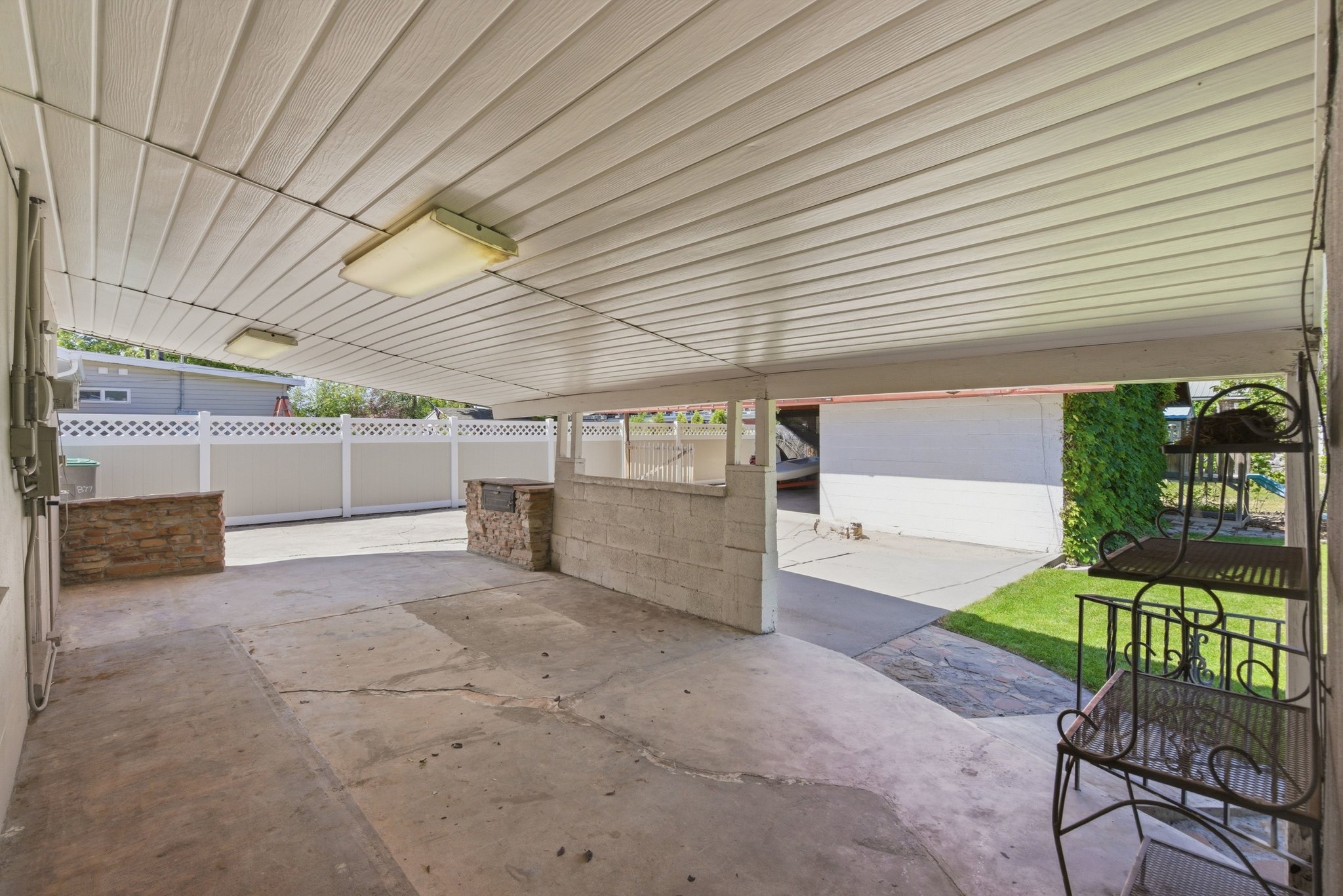 Covered outdoor concrete patio with overhead lighting, stone-clad retaining walls, and a white lattice fence