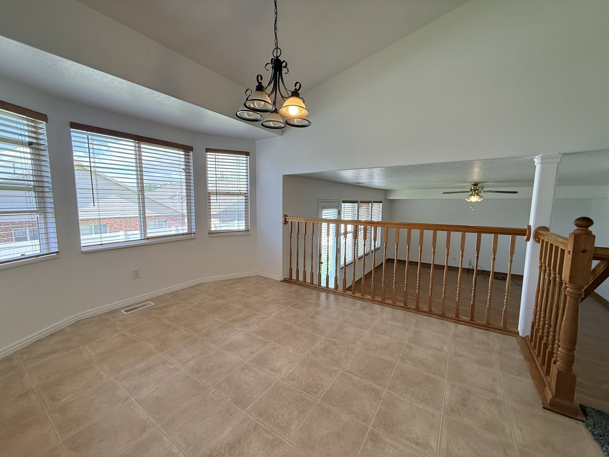 Vaulted ceiling with a chandelier light fixture, multiple windows with blinds, tiled flooring, wood railing, and a view to the lower level with wood-finish flooring