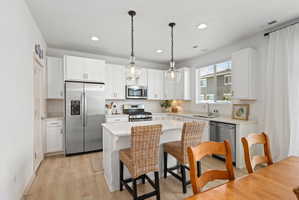 Bright kitchen featuring white cabinetry, stainless steel appliances, a center island with countertop seating, wood-finish flooring, and pendant lighting