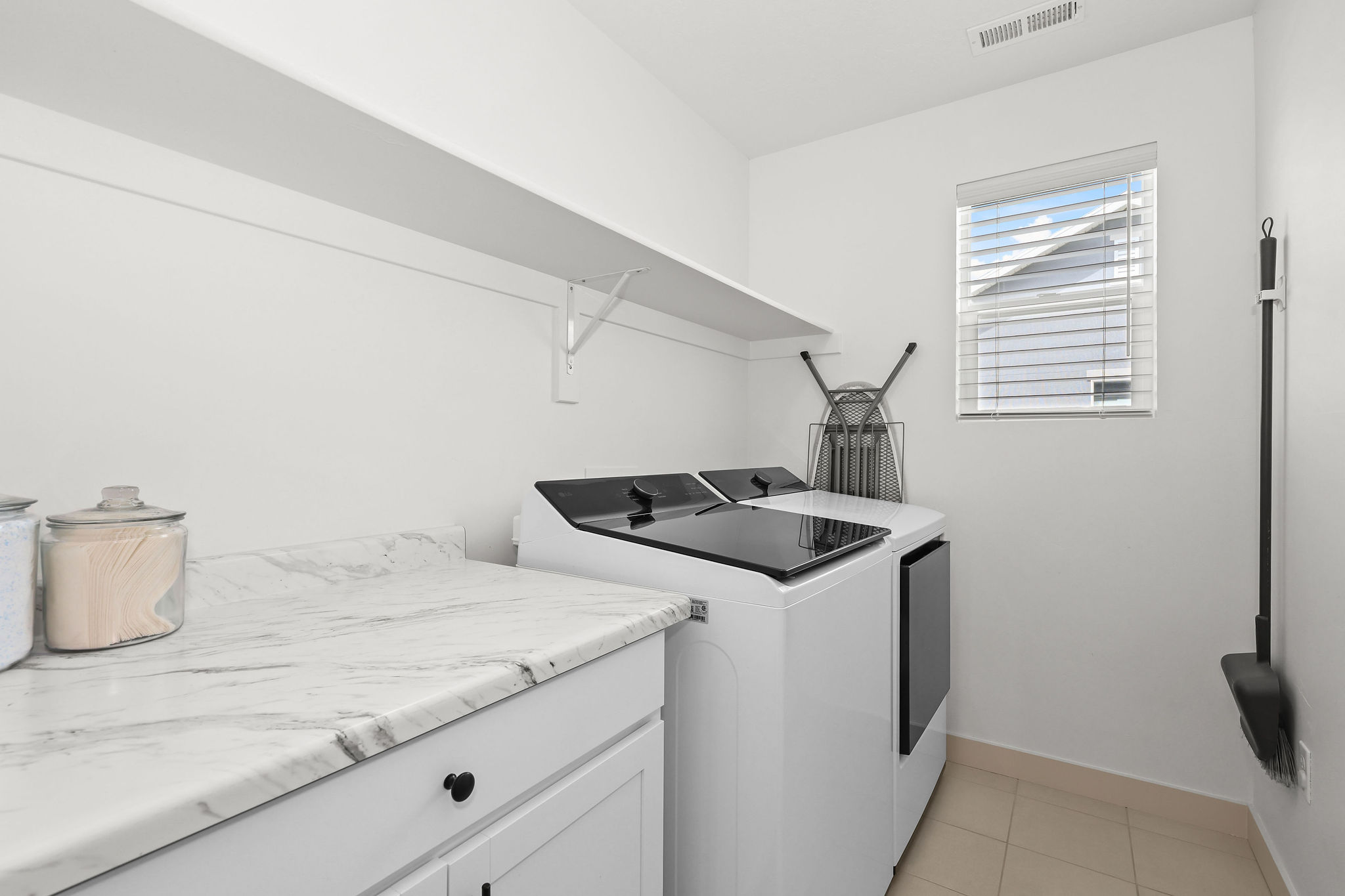 Dedicated laundry space featuring white cabinetry with black hardware, a light-toned countertop with veining, an upper storage shelf, and light tile flooring