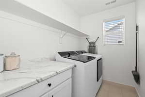Dedicated laundry space featuring white cabinetry with black hardware, a light-toned countertop with veining, an upper storage shelf, and light tile flooring