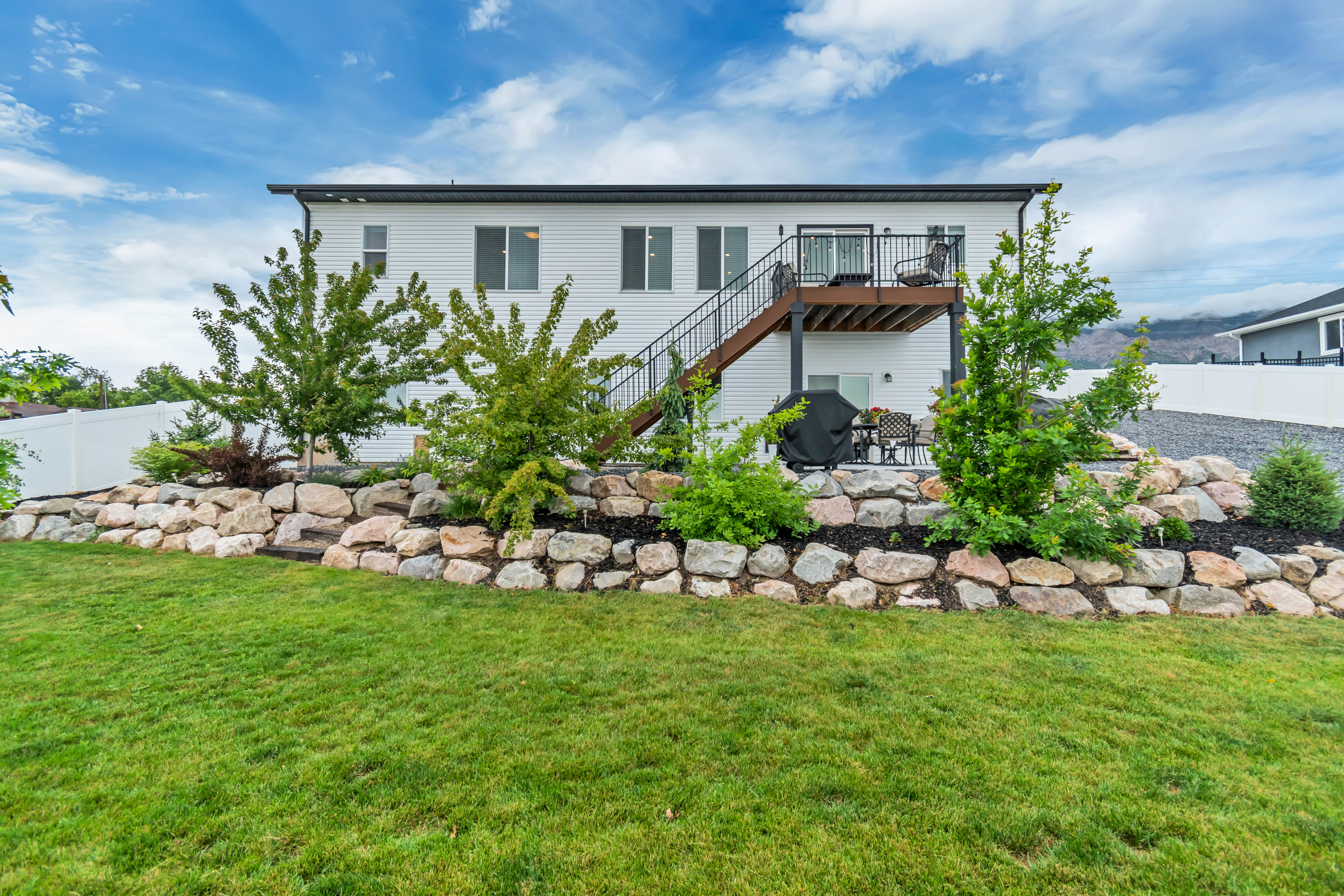 Rear exterior featuring white siding, a raised deck with dark railing and stairs, a lower patio, and tiered landscaping with natural rock retaining walls and plantings