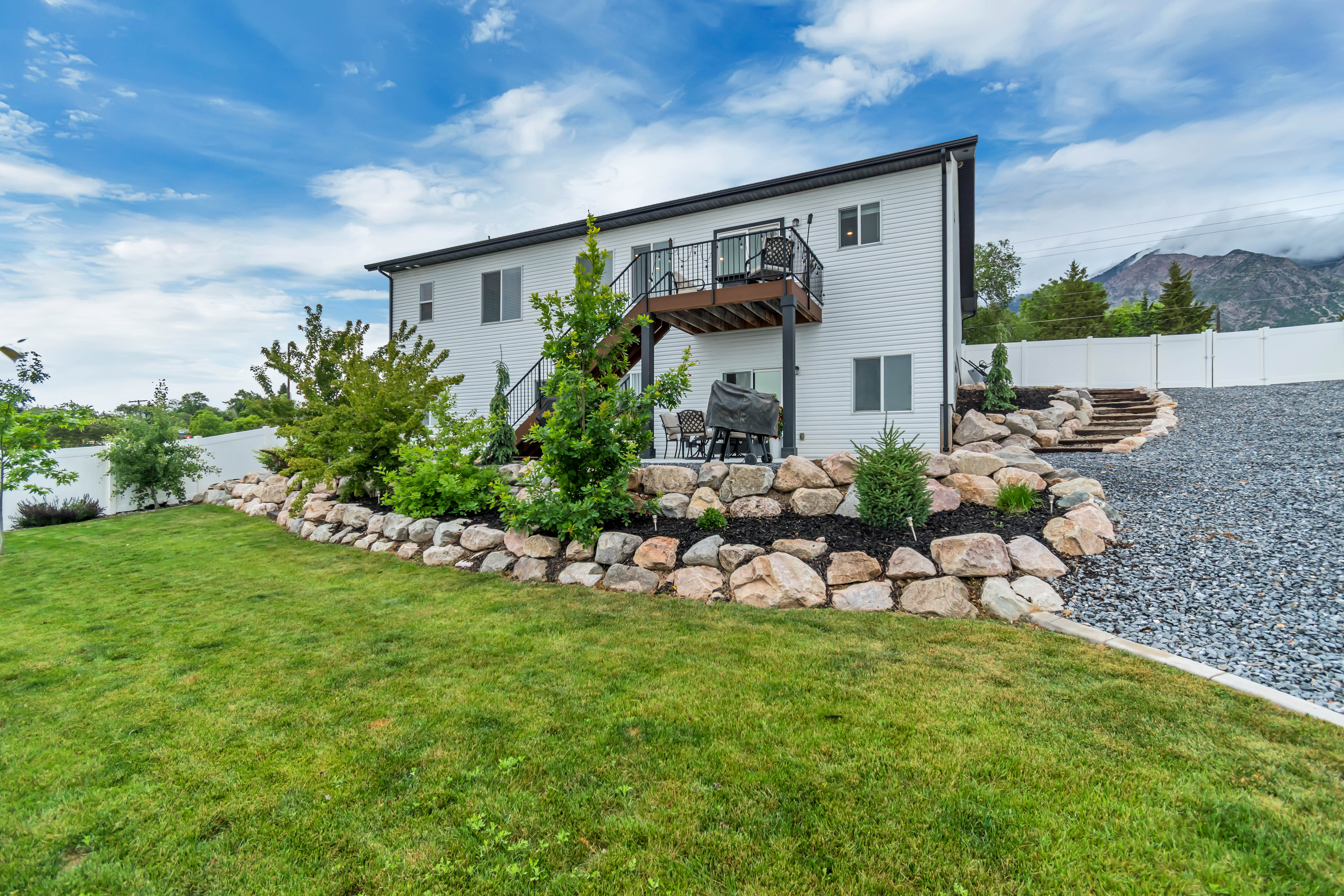 Expansive backyard featuring a manicured lawn, a rock retaining wall with landscaping, a gravel path with stone steps, a white privacy fence, and a mountain backdrop
