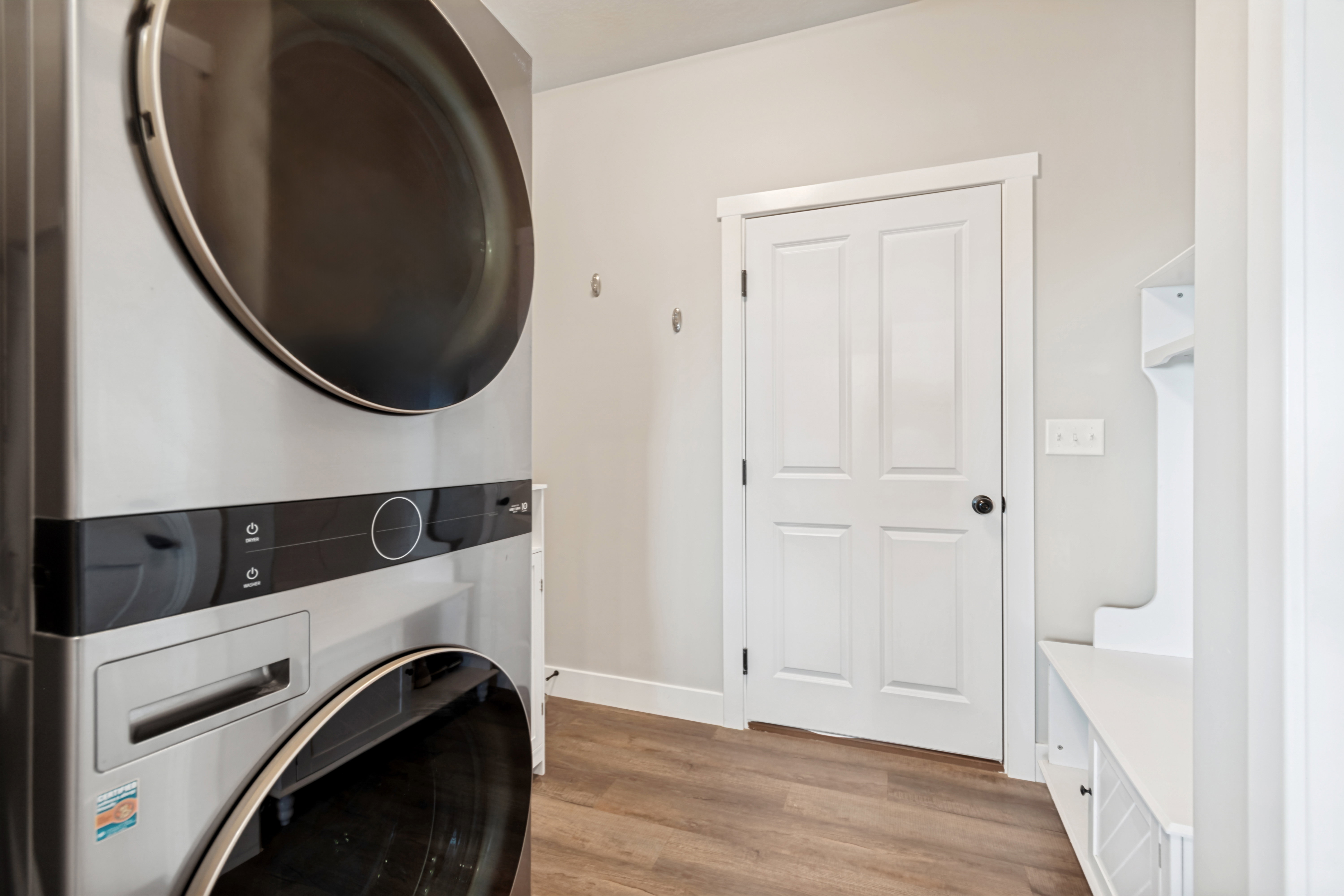 Integrated laundry system with a stacked washer and dryer, wood-finish flooring, white paneled door with black hardware, and built-in white bench seating