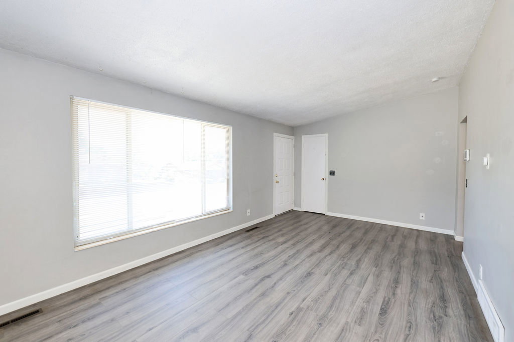 Spacious living area featuring light gray walls, wood-finish flooring, a large window with blinds, and white trim throughout