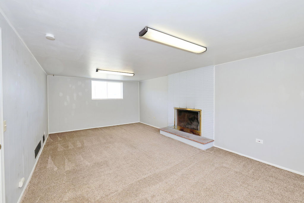 Spacious room featuring neutral carpet flooring, a white painted brick fireplace with a stone hearth, and a window providing natural light