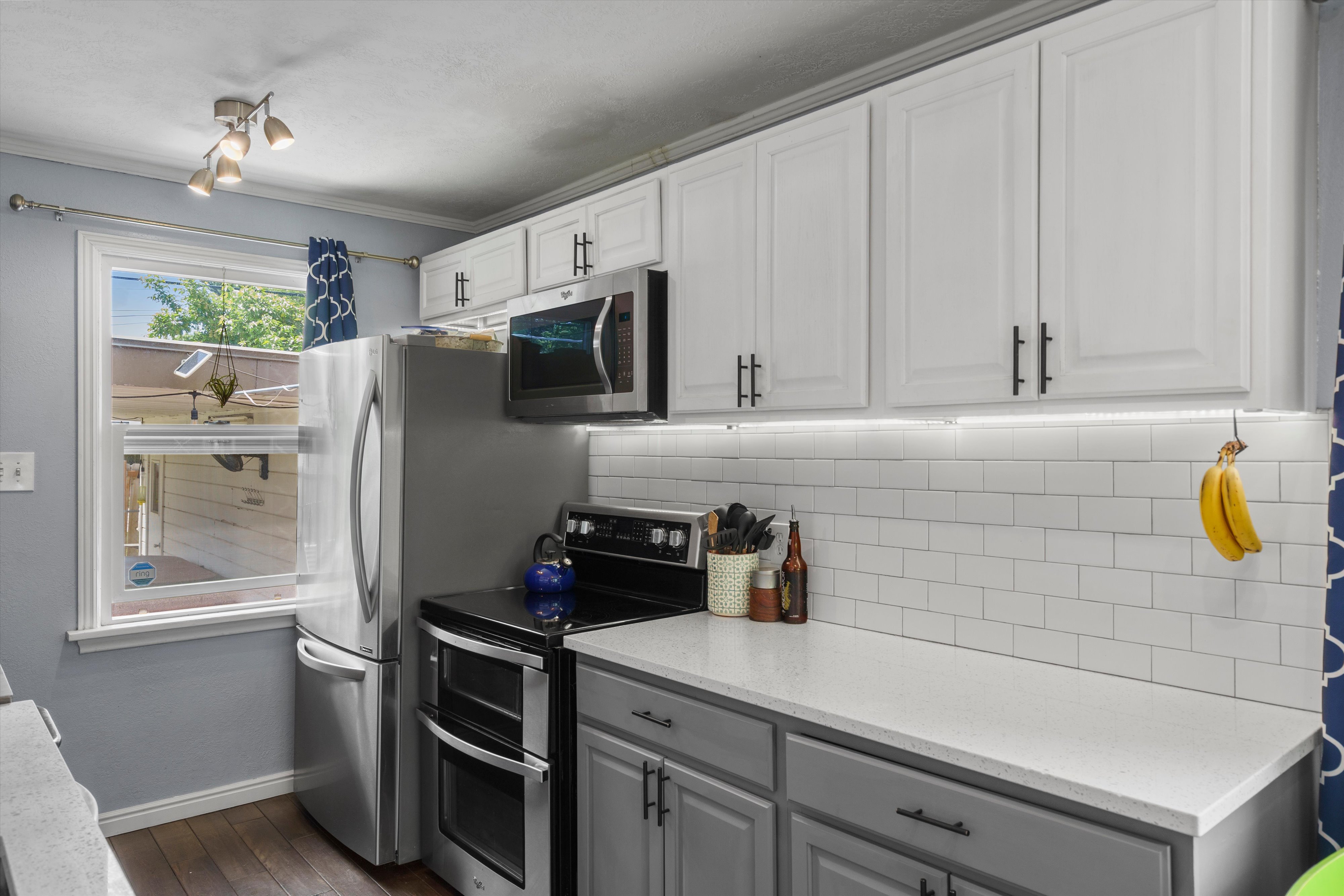 Bright kitchen featuring two-tone cabinetry with modern hardware, stainless steel appliances, a subway tile backsplash, under-cabinet lighting, and light-toned countertops