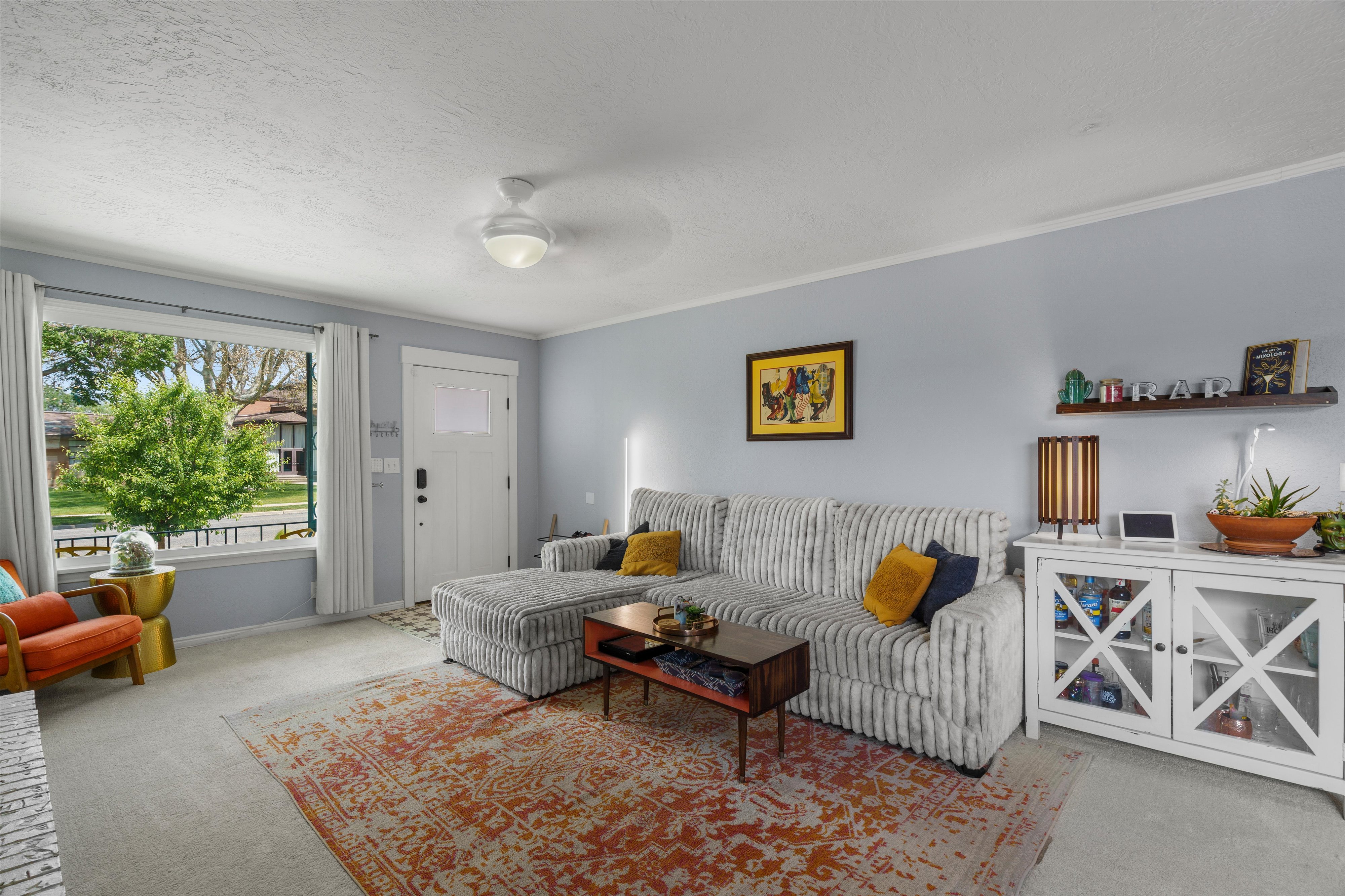 Bright living area featuring a large picture window, light blue wall paint, a white front entry door with a rectangular glass insert, and a ceiling-mounted light fixture