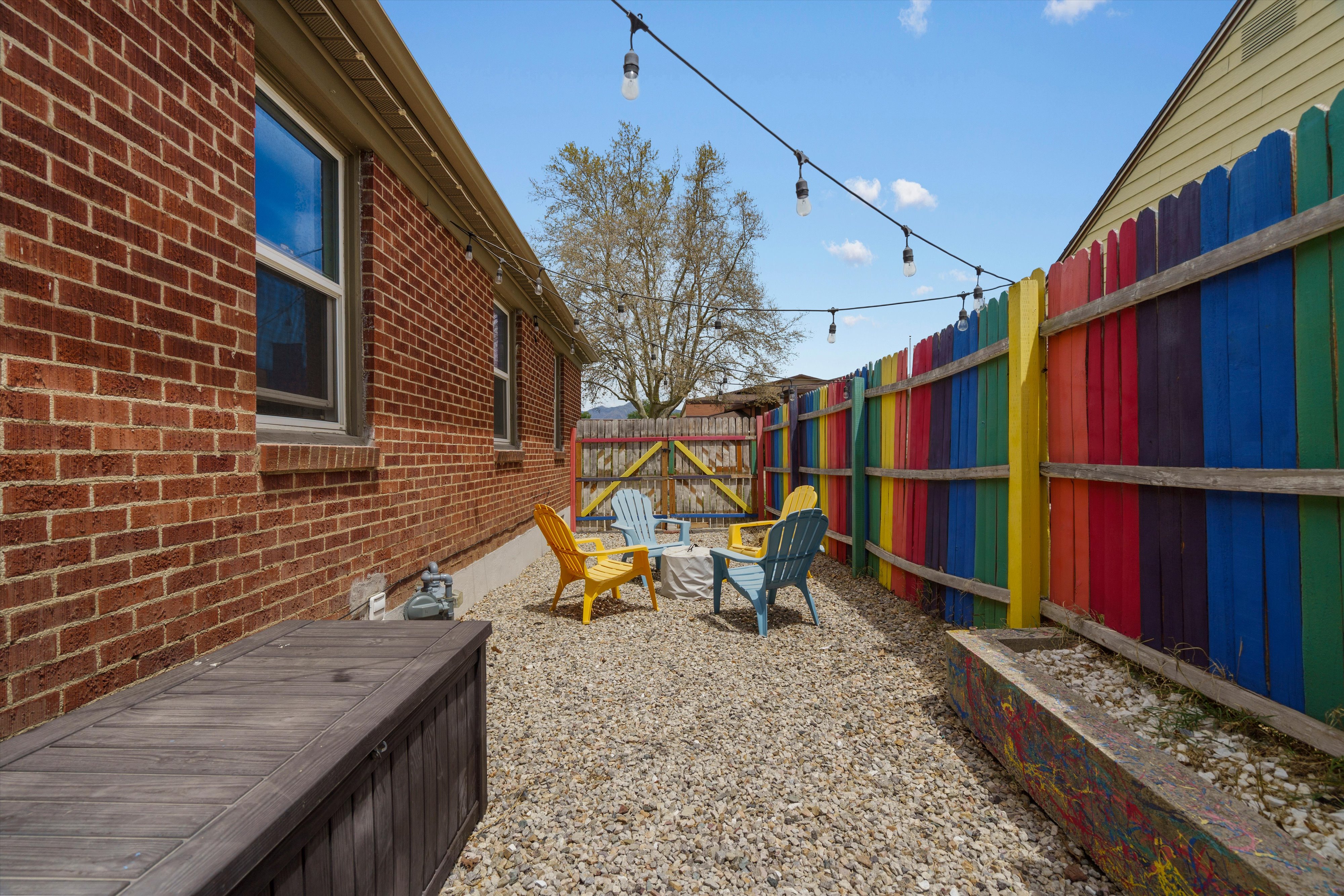 Gravel-paved outdoor space featuring string lighting, a colorful privacy fence, and a brick exterior wall