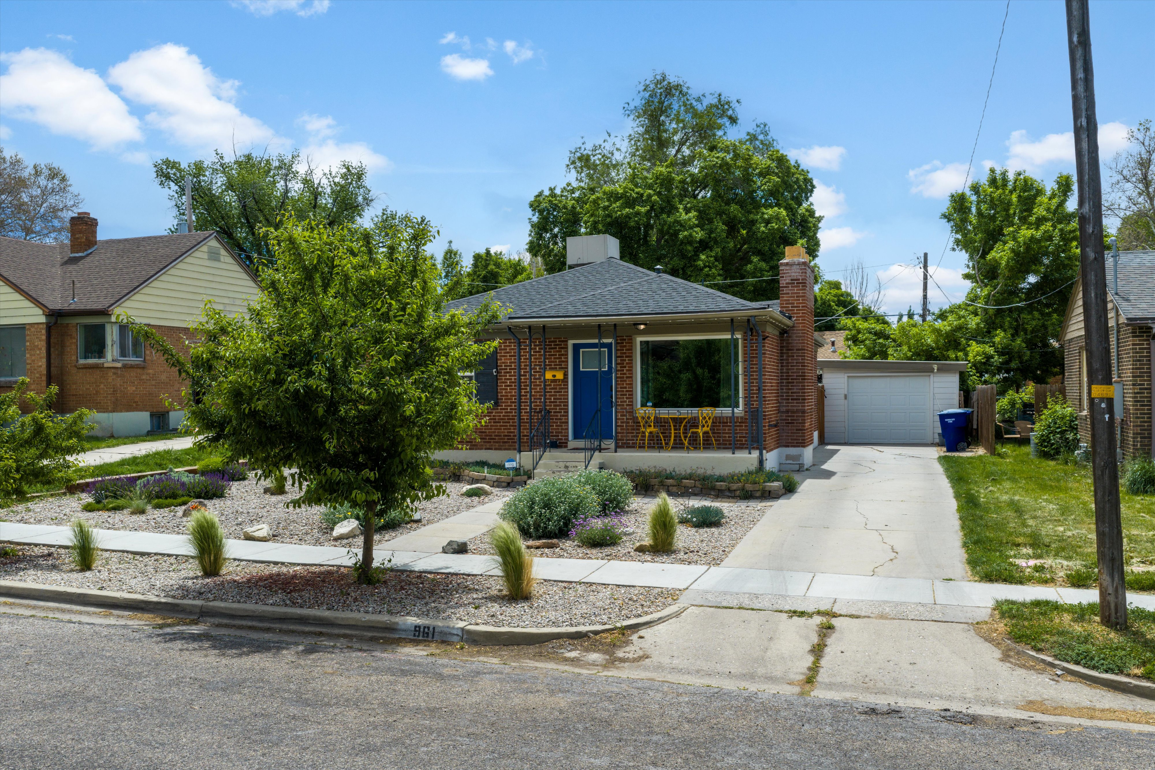 Brick facade residence featuring a prominent picture window, a bright blue front door, a concrete driveway, a detached garage, and a low-maintenance xeriscape front yard with mature plantings