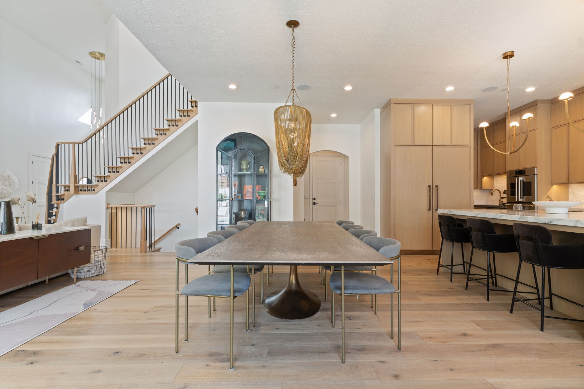 Open-concept living area featuring light wood-finish flooring, a contemporary dining area with a distinctive chandelier, a gourmet kitchen with Monogram appliances and a large island, and a prominent staircase with dark balusters